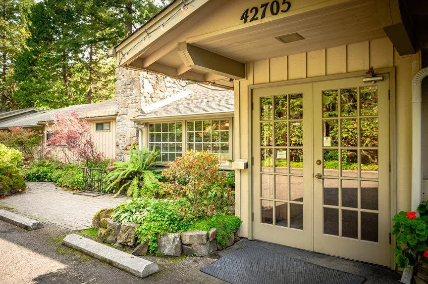 Exterior view of a residential building with a glass double door, surrounded by greenery and flowering plants, and a stone pathway leading to the entrance.