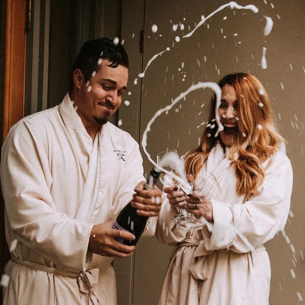 Two people in bathrobes celebrating with champagne and popping a bottle, with foam and spray flying in the air.