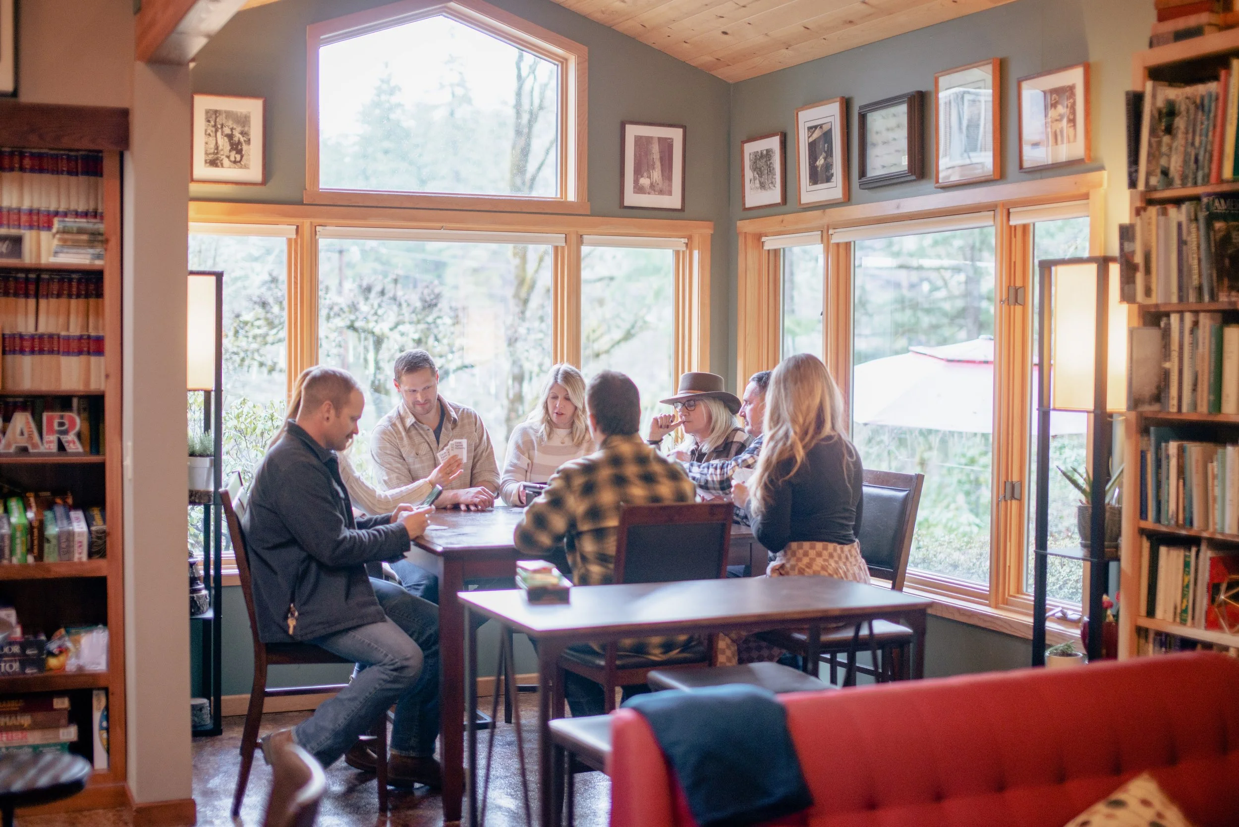 Group of people sitting around a table playing a card game in a cozy, well-lit room with large windows and bookshelves.