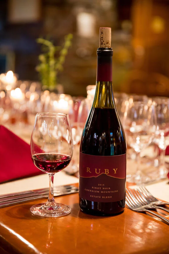 A bottle of Ruby 2016 estate blend wine with a glass of red wine on a wooden table at a dining setting with a red napkin, silverware, and blurred candlelit background