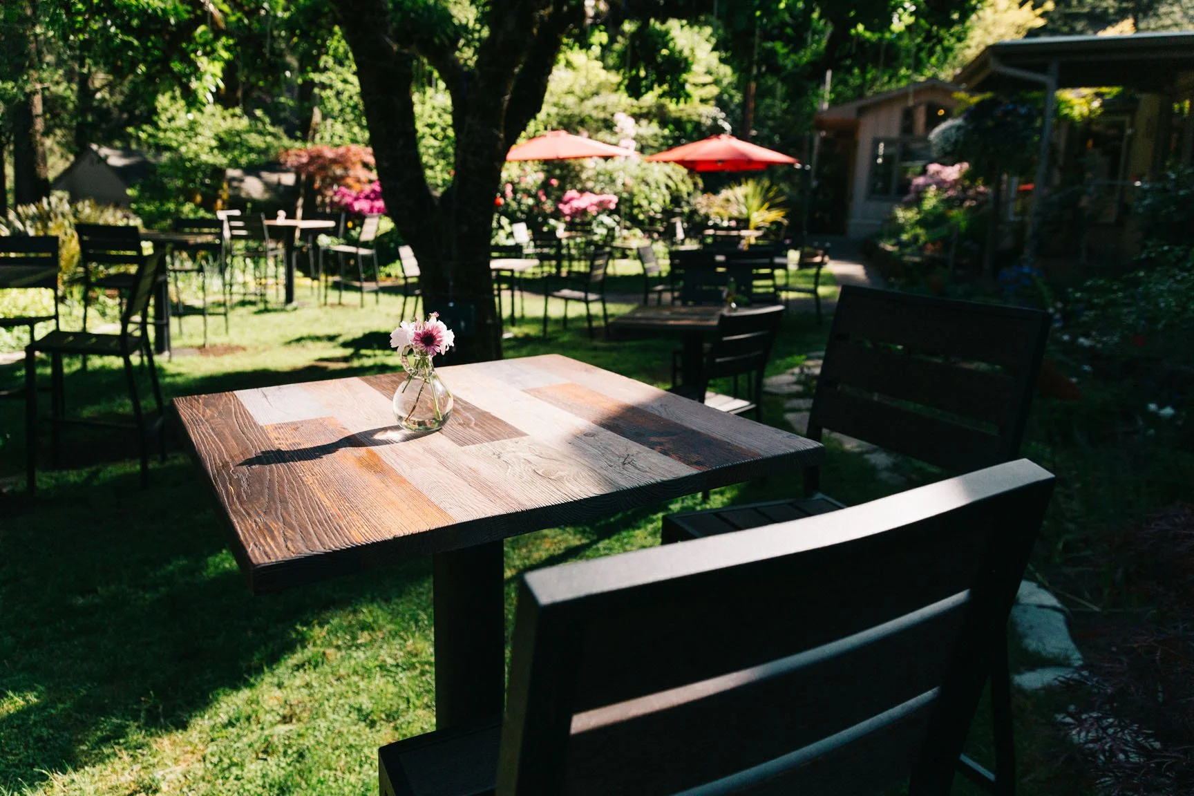 A wooden table with a small vase of pink and white flowers on a sunny outdoor patio surrounded by empty black chairs and tables, with trees, shrubs, and red umbrellas in the background.