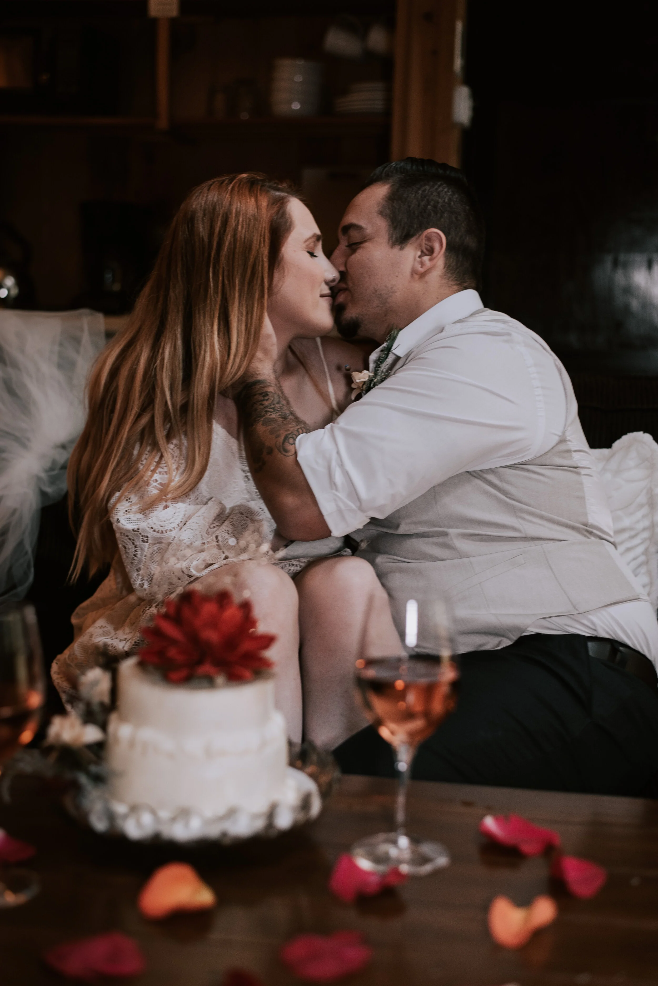 A couple shares an intimate moment at a wedding celebration, seated close with their faces nearly touching, in front of a wedding cake topped with a red flower and glasses of wine. Rose petals are scattered on the table.