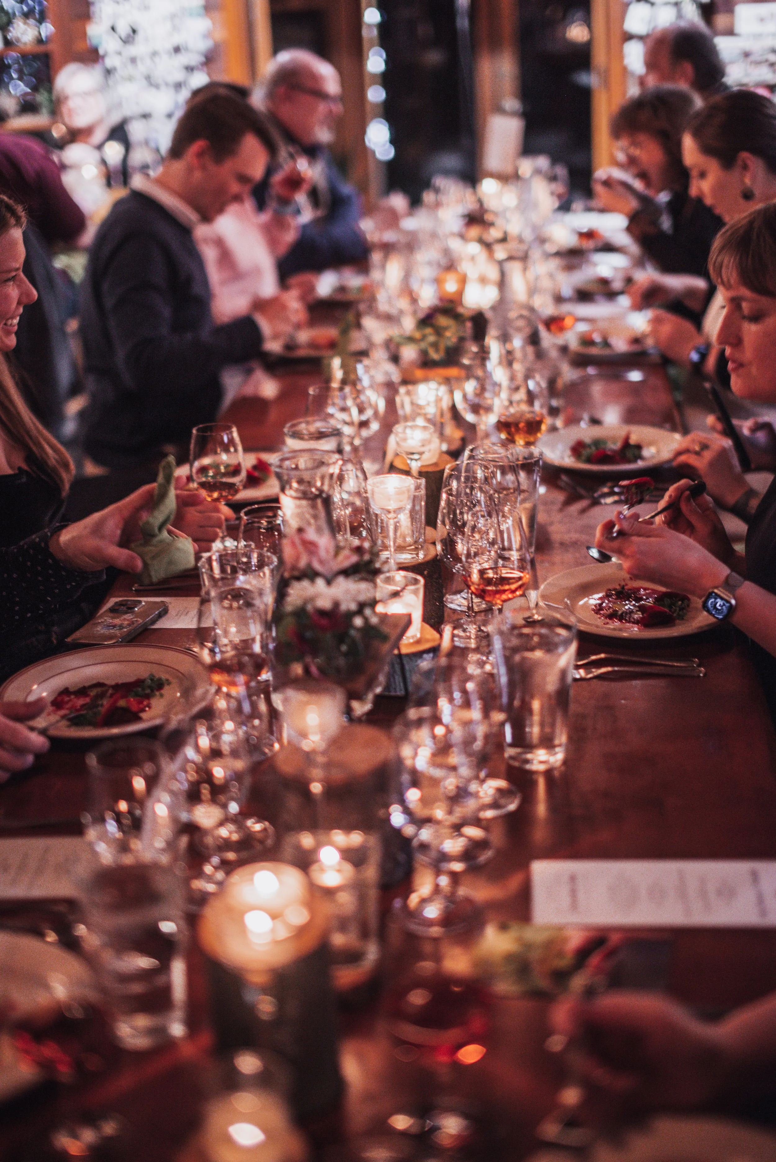 A group of people dining at a long, candle-lit table, enjoying a meal with plates, glasses of wine, and decorative centerpieces.