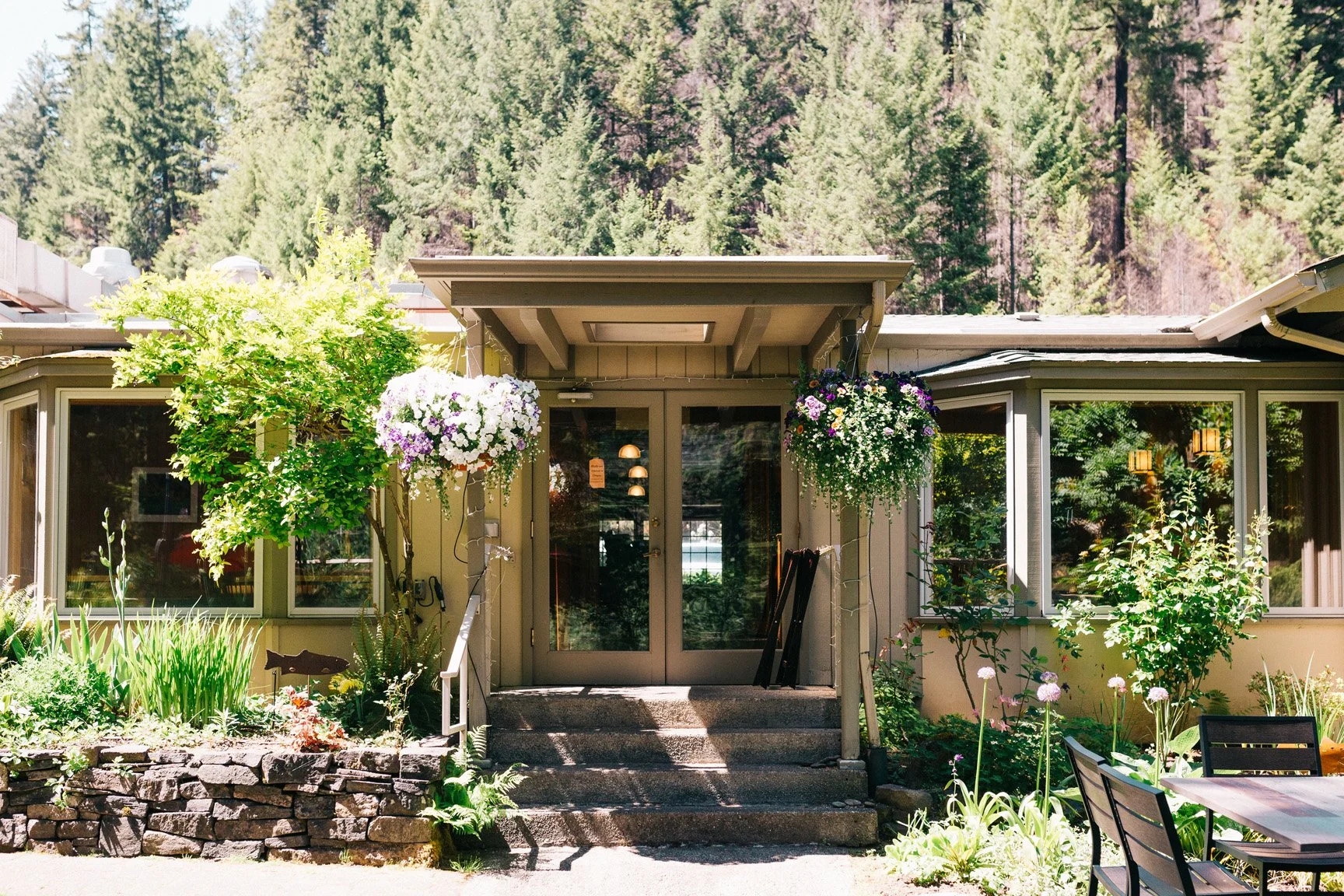Front entrance of a house with glass double doors, surrounded by plants and hanging flower baskets, with a mountain forest background.