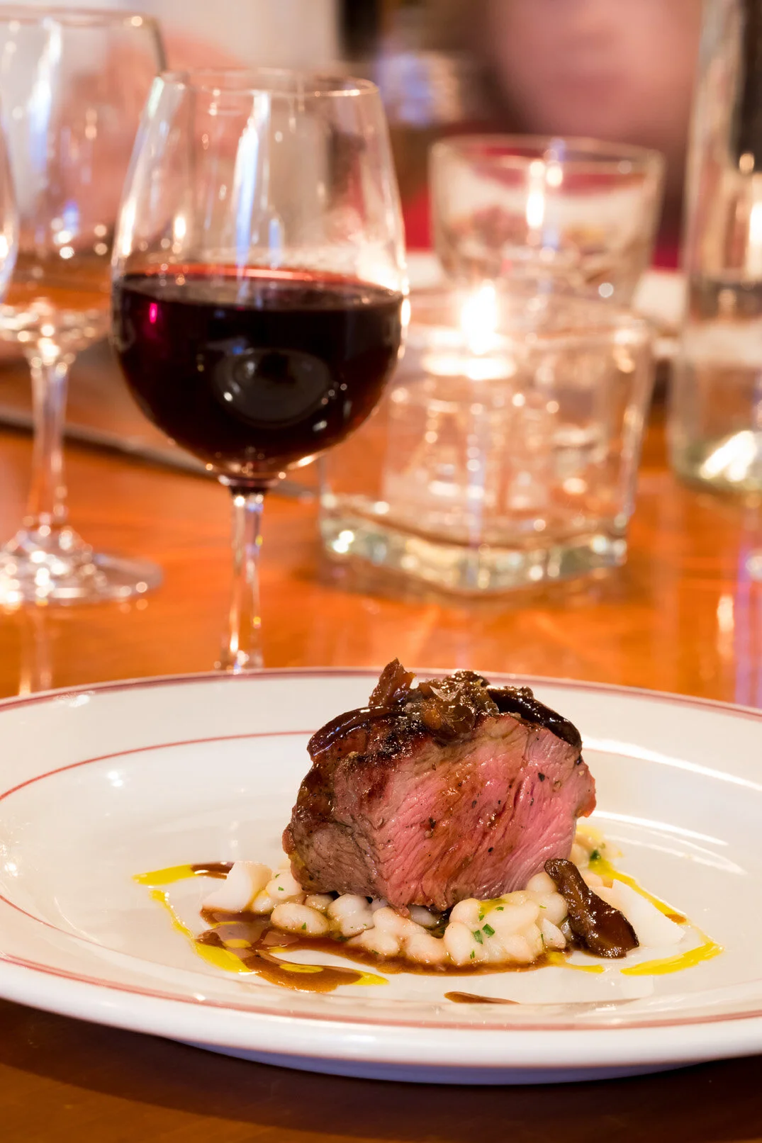 A gourmet dinner plate with a cooked steak sitting on a bed of white beans, drizzled with olive oil and balsamic reduction, with a glass of red wine in the background and a candle on a wooden table.
