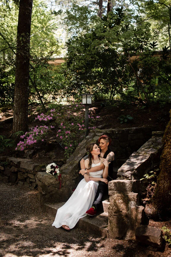 Brides sitting on stone steps in a garden with greenery, pink flowers, and a lamp post, dressed in wedding attire.