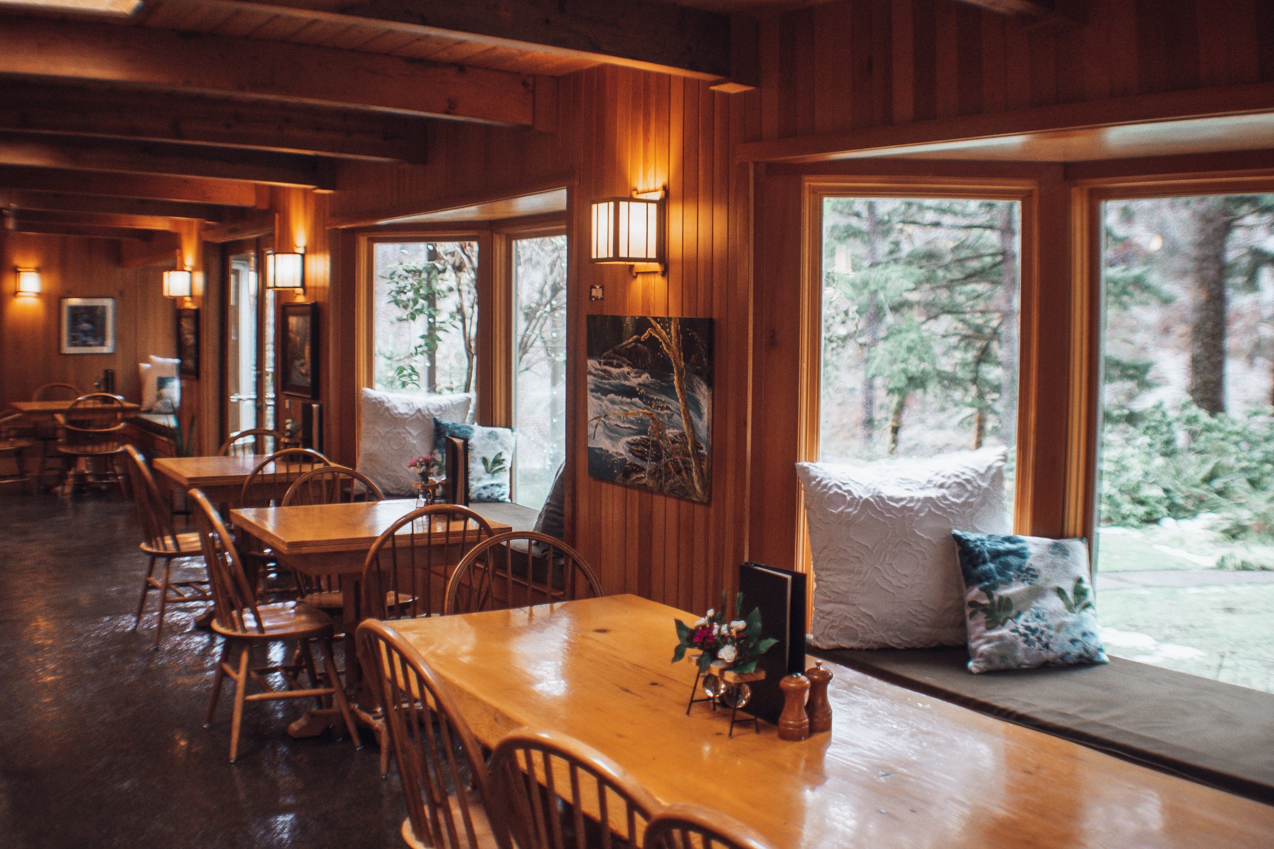Cozy wooden interior of a restaurant with large windows, wooden tables, and chairs, decorated with cushions and small floral arrangements.