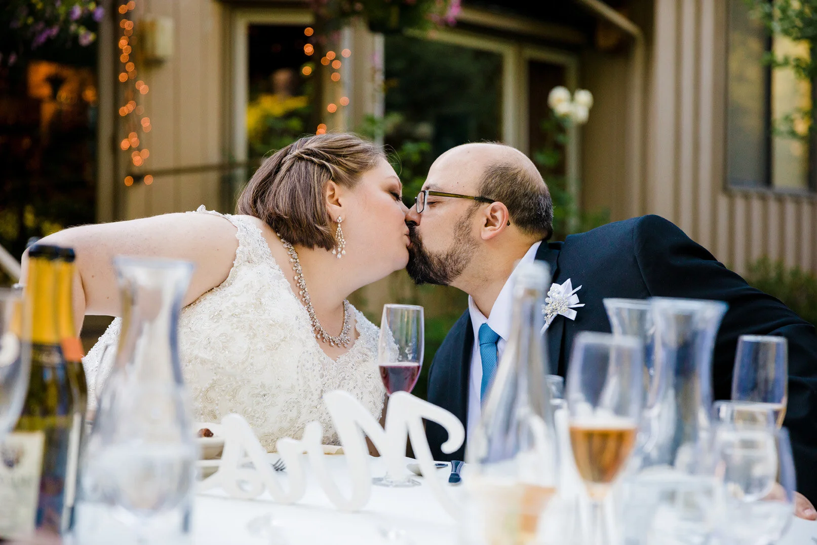 A newlywed couple sharing a kiss at their wedding reception, seated at a table with glasses and decorative items, outdoors with string lights and a building in the background.