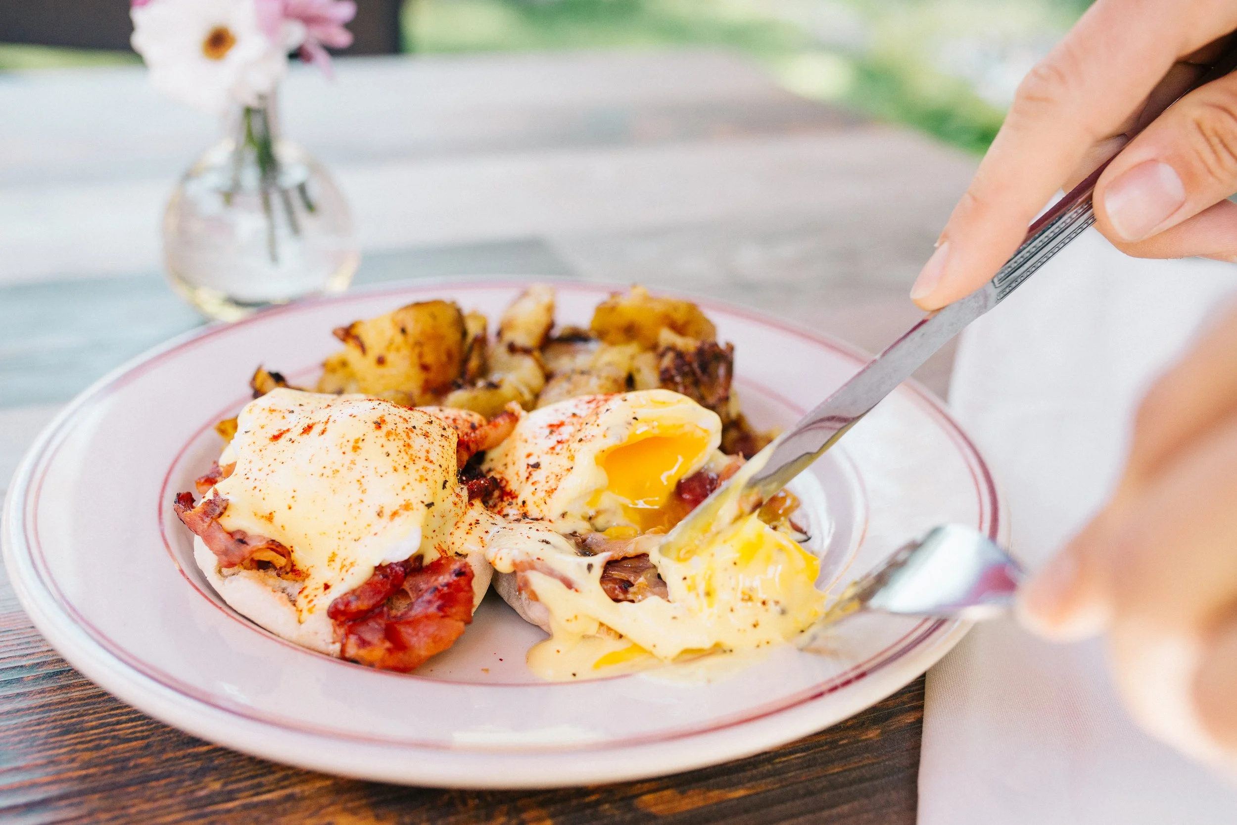 Close-up of a plate with eggs Benedict, potatoes, and bacon, with someone cutting into the eggs. In the background, there is a small vase with pink and white flowers.
