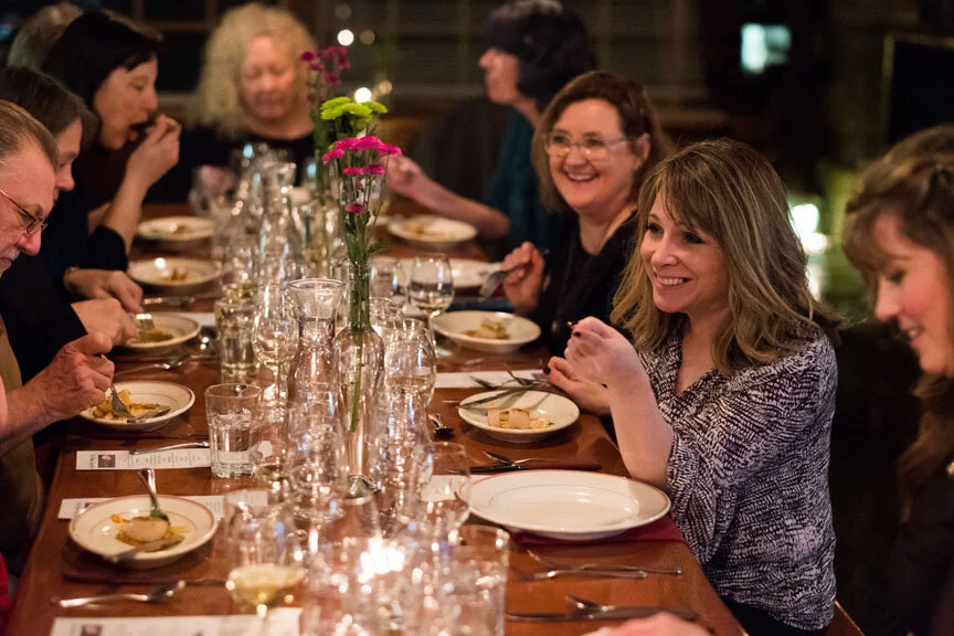 People sitting at a dining table, laughing and engaging in conversation, with flower arrangements and plates of food.
