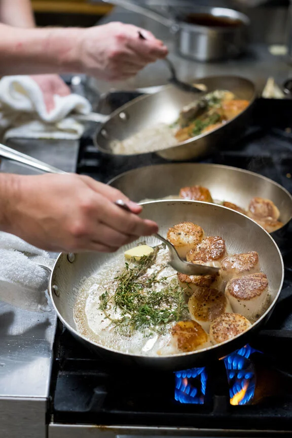 Chef cooking scallops with herbs and garlic in a frying pan on a stovetop.