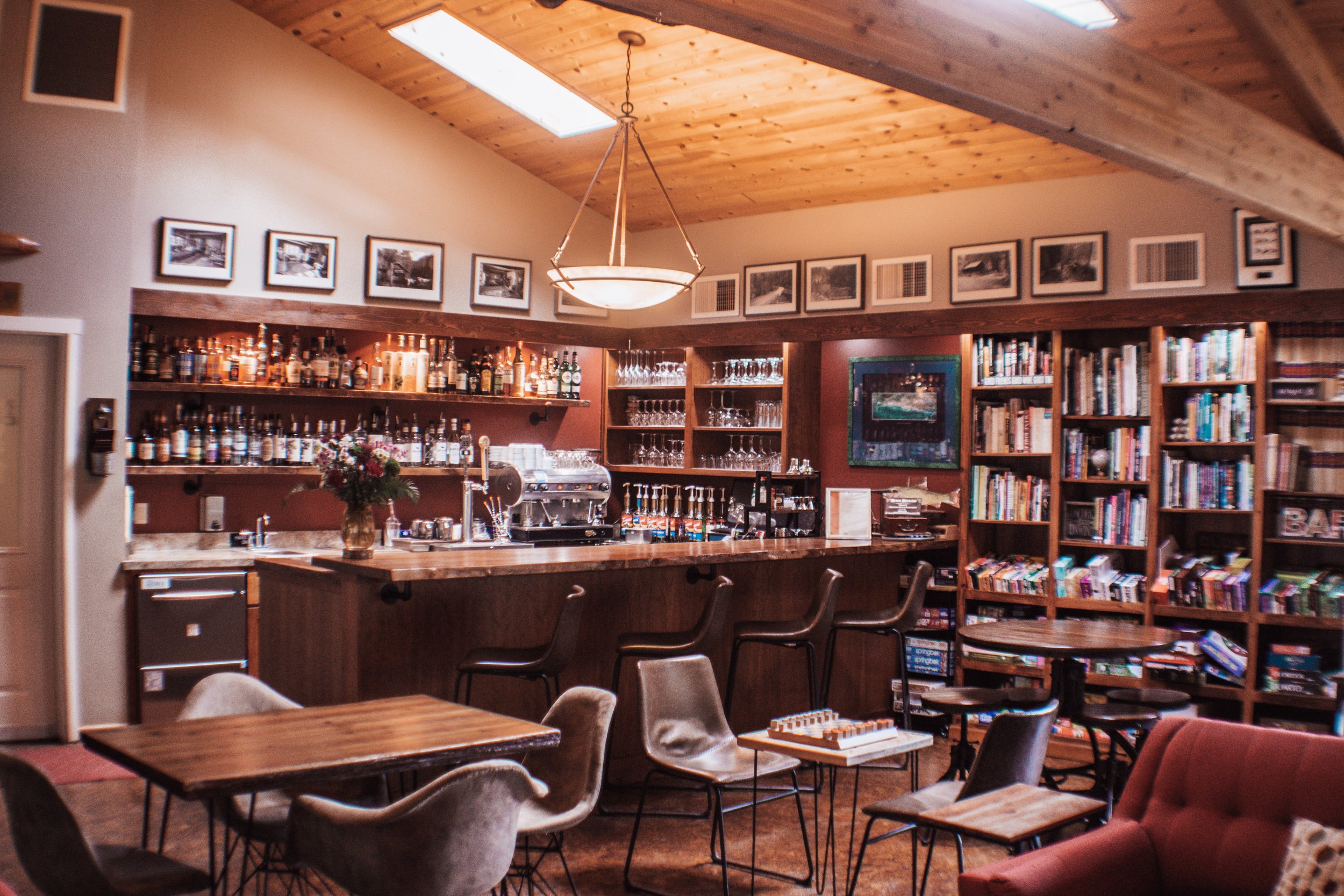 Cozy cafe or lounge area with a bar counter, bookshelves, and seating areas with chairs and a couch, under a wooden ceiling with skylights.