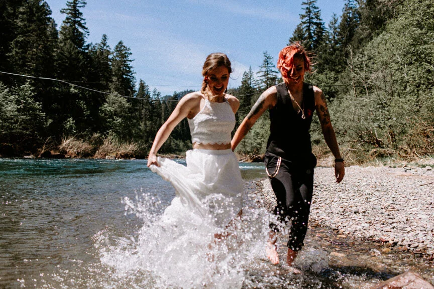Two women walking barefoot in the water at a riverbank, smiling and holding hands, surrounded by forested mountains under a clear sky.