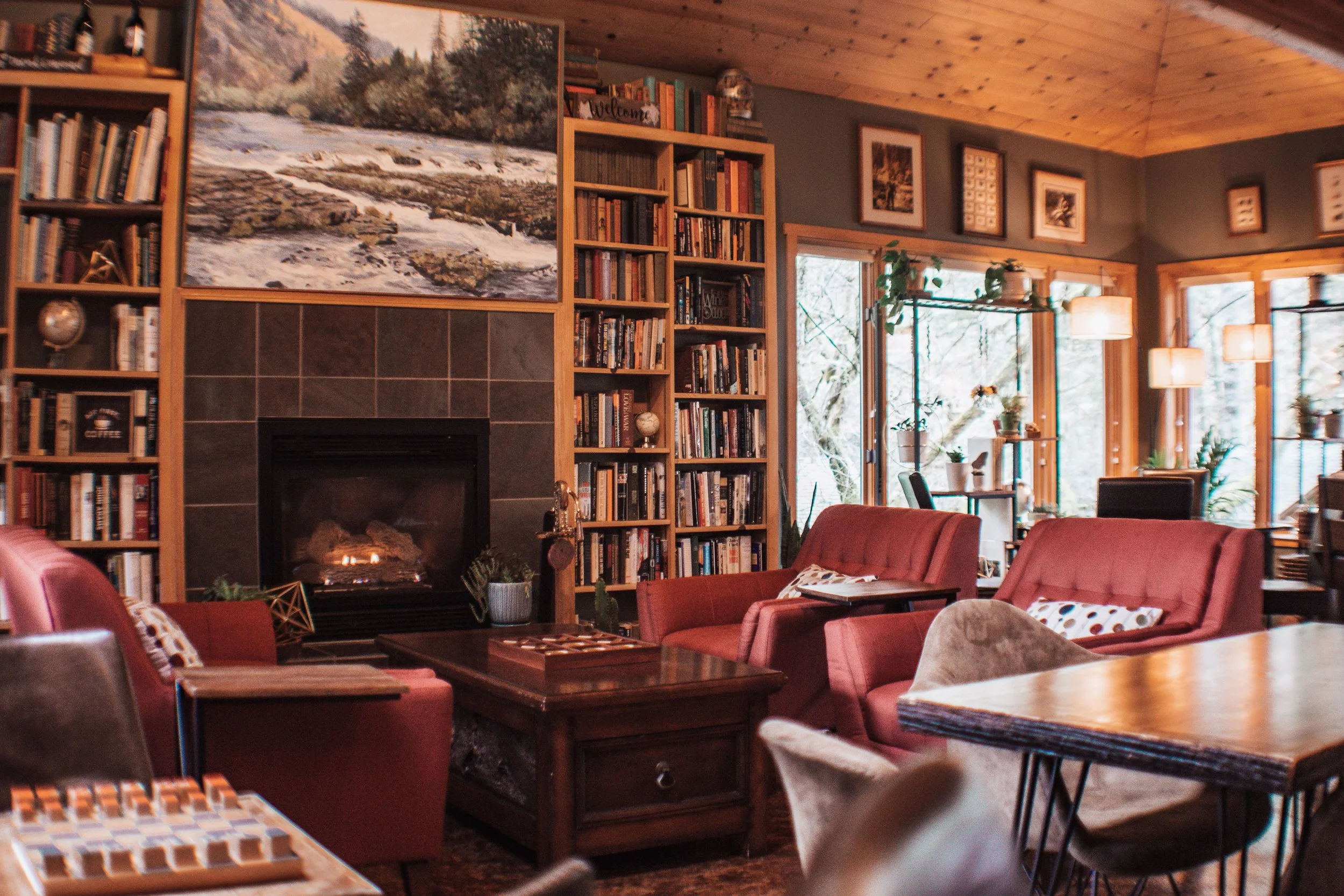 Cozy living room with a fireplace, wooden bookshelves filled with books, red armchairs, a coffee table, and large windows with plants and hanging lights.