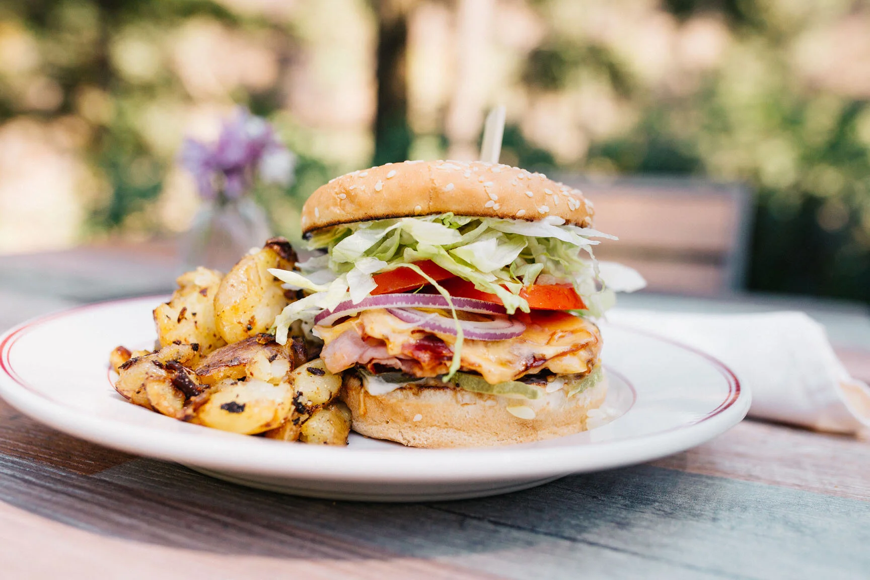 A plate with a cheeseburger topped with lettuce, tomato, onions, and bacon, served with seasoned roasted potatoes, outdoors with a blurred background.