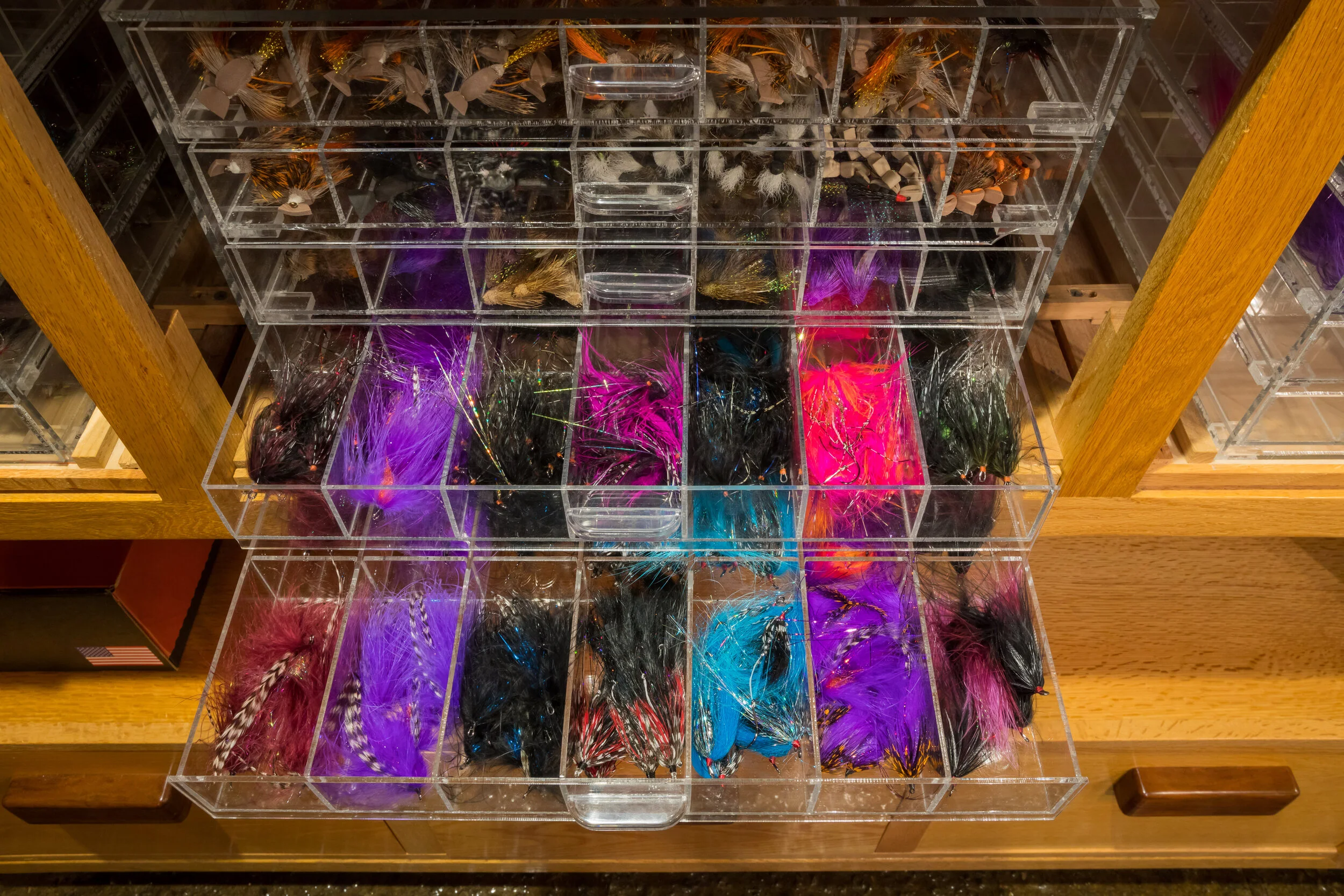 A wooden display case containing multiple clear plastic compartments, each filled with colorful fishing lures and flies, organized by type and color.