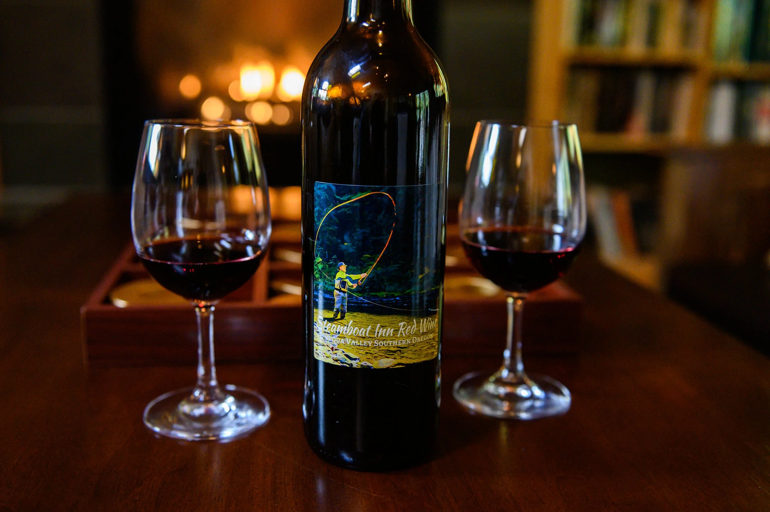 A bottle of red wine with a label depicting a fisherman casting a line, flanked by two glasses of wine, set on a wooden table in front of a fireplace and a bookshelf.