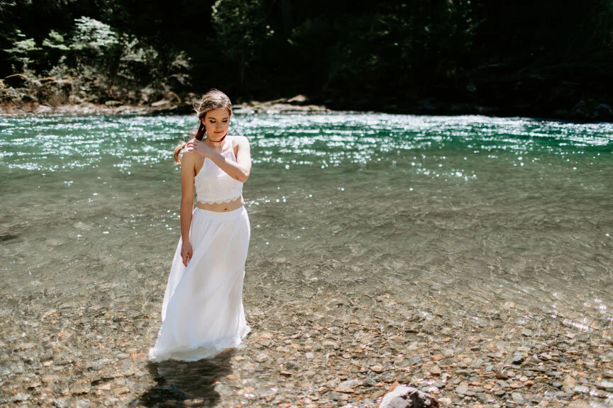 A woman in a white dress standing in a clear river with a rocky riverbed, surrounded by trees and lush greenery.