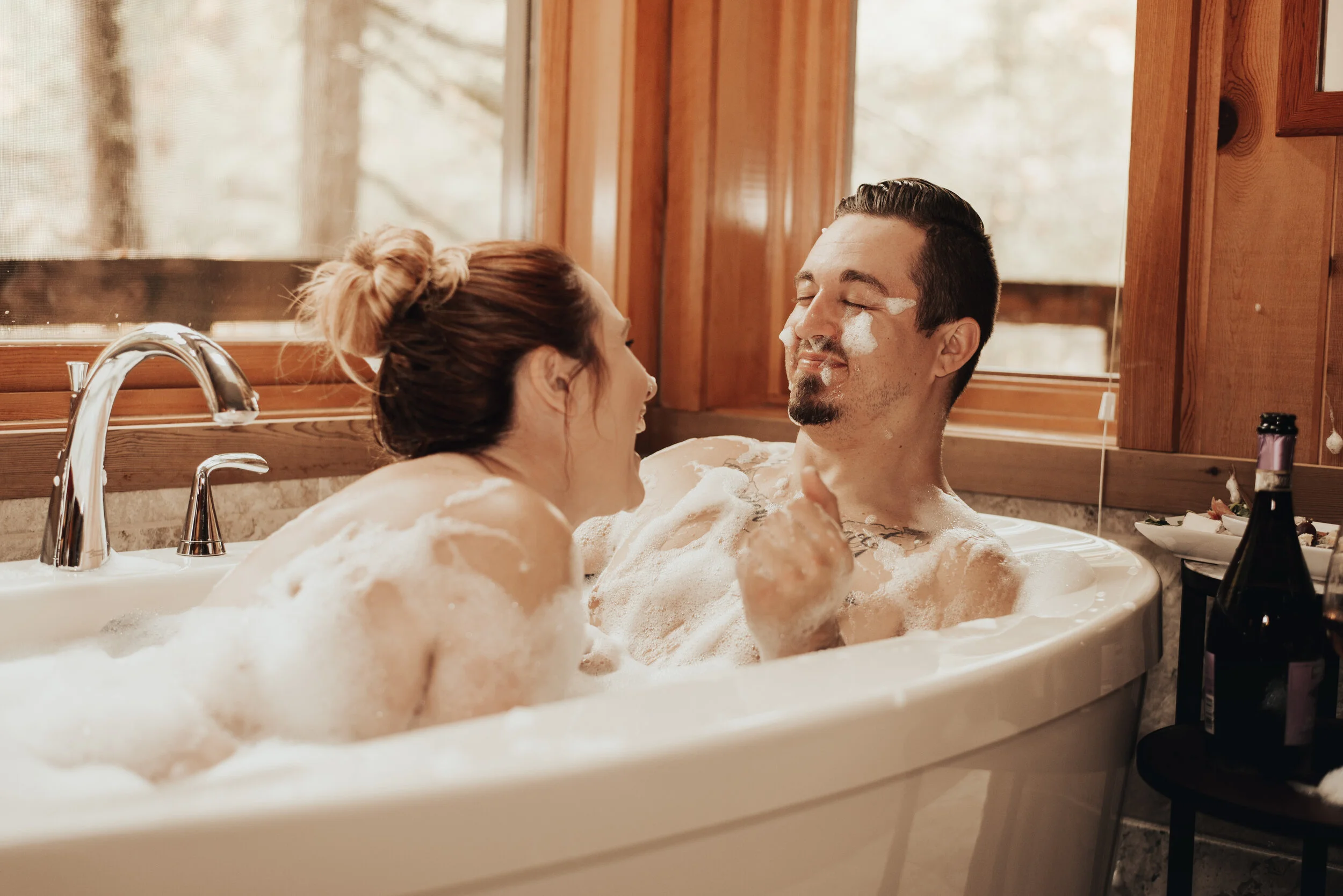 A couple enjoying a bubble bath together in a bathroom with wood walls and a large window, with soap, bubbles, and a bottle of wine nearby.