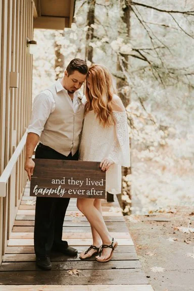A couple stands close together on a wooden porch, holding a sign that reads 'and they lived happily ever after'. The woman has long red hair and wears a white dress, while the man has dark hair and is dressed in a white shirt and dark pants. They are