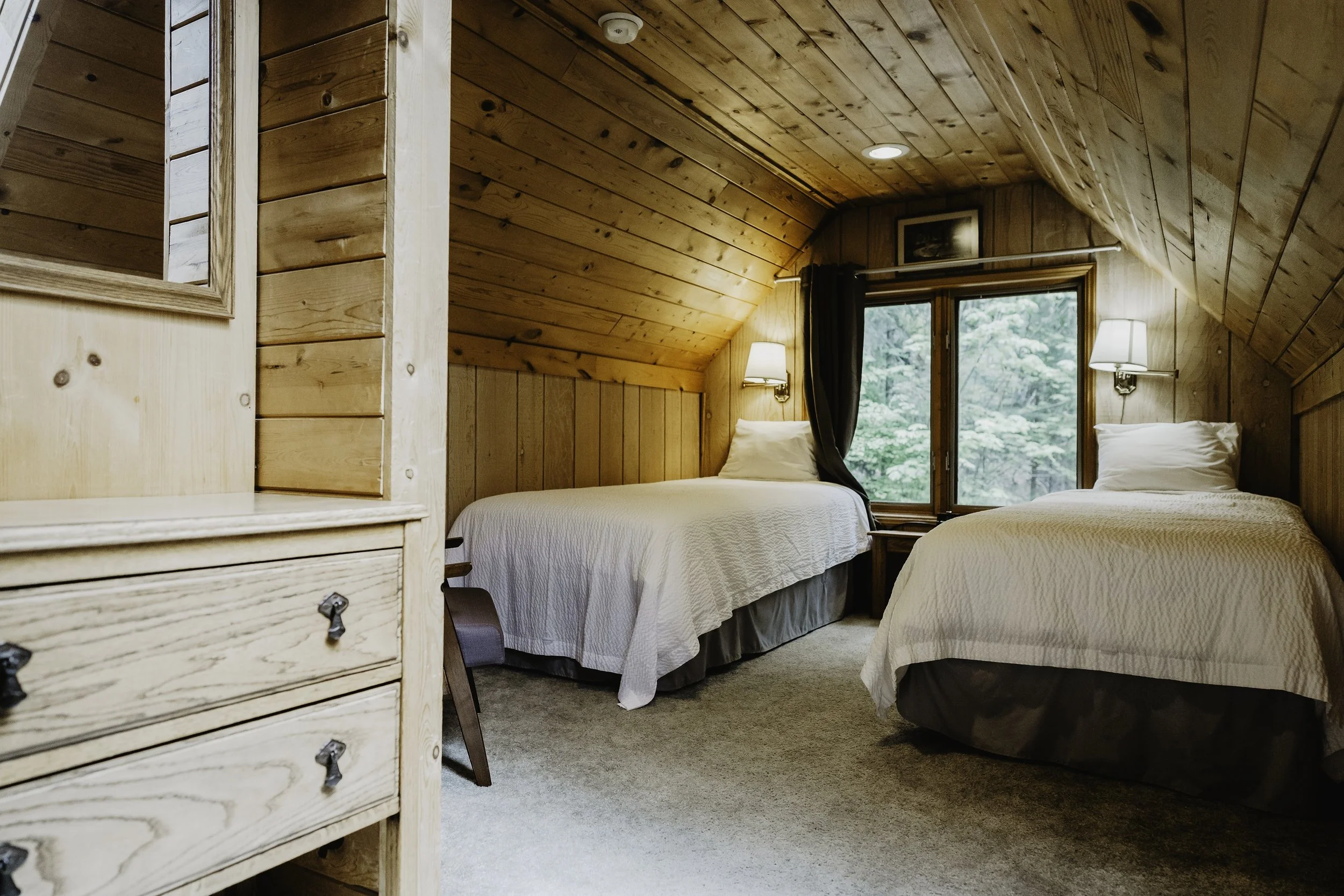 A cozy bedroom with wood-paneled walls and ceiling, featuring two twin beds with white bedding, a window with curtains showing greenery outside, and wall-mounted lamps.