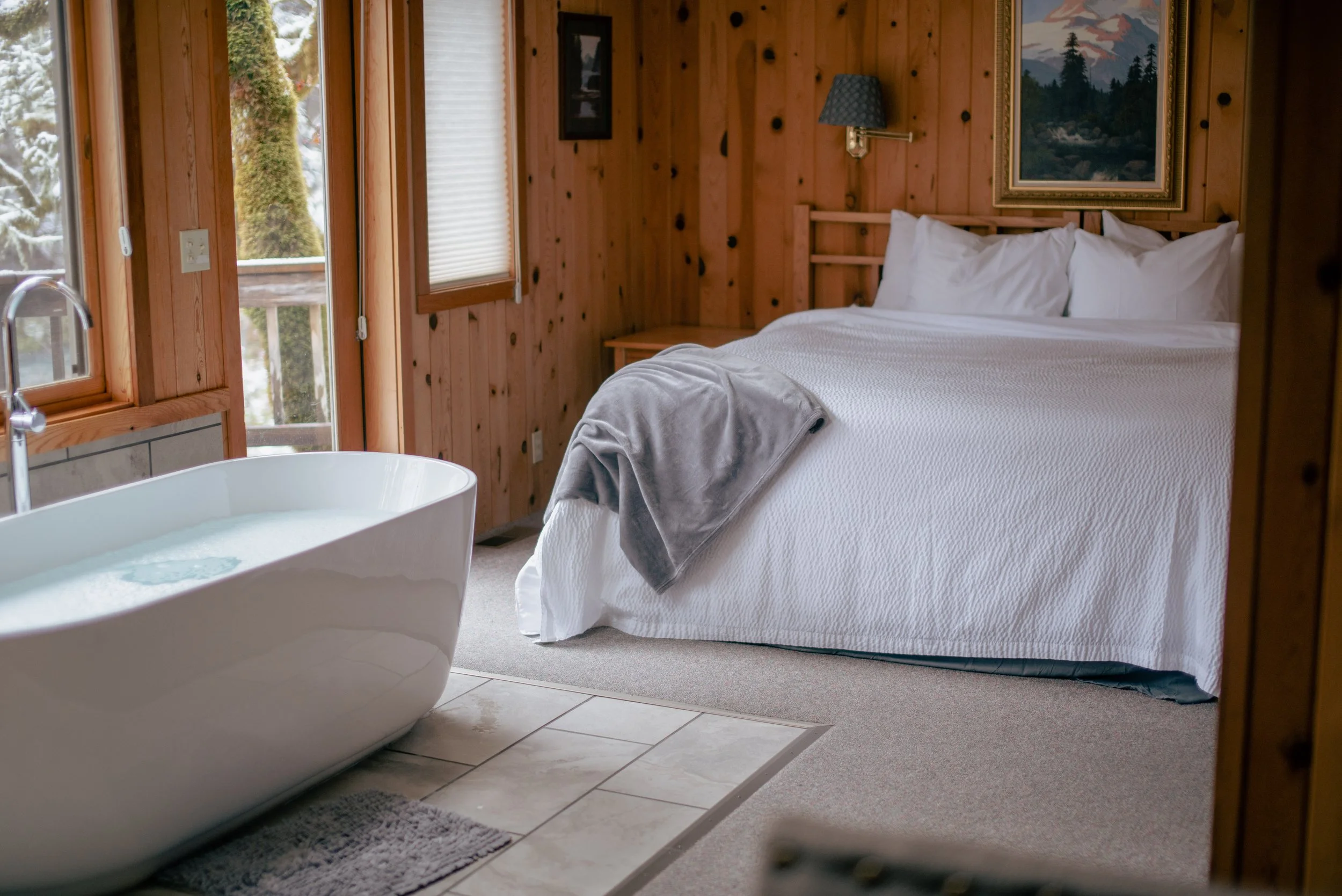 A rustic hotel room with wooden walls, a large bed with white bedding and pillows, a gray blanket draped over the side, a small bedside table, a lamp, framed artwork on the wall, and a freestanding bathtub in front of a window showing outdoor trees.