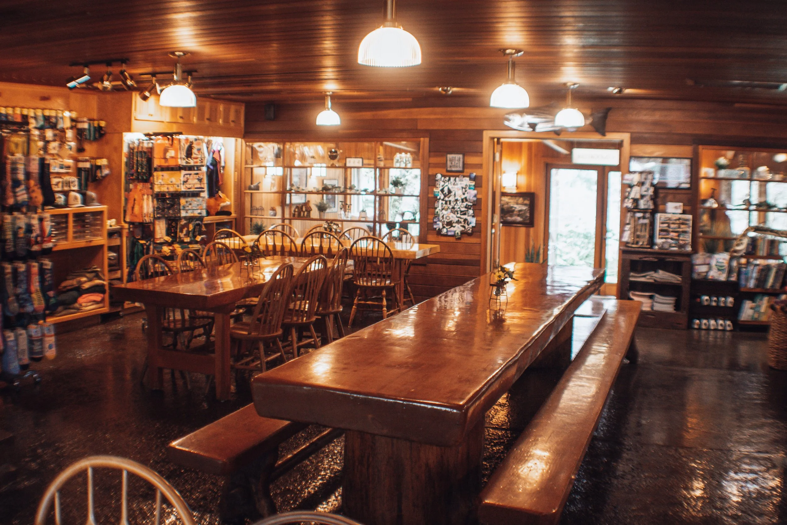 A cozy interior of a wooden store or cafe with long wooden tables, chairs, and shelves filled with various items and books, warm lighting, and a door leading outside.