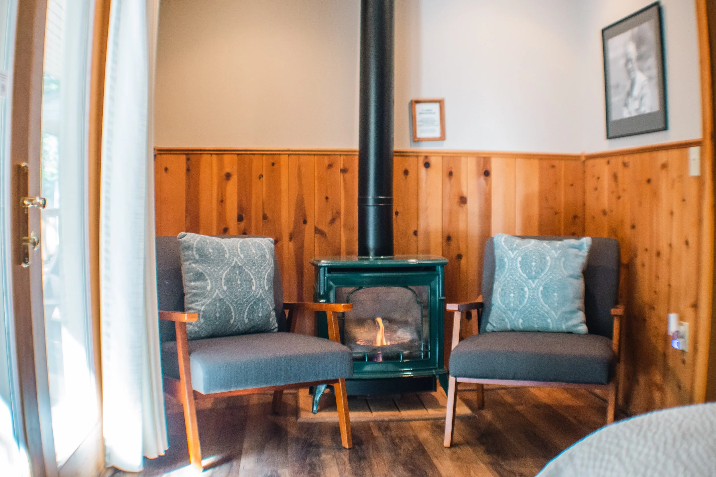 Two gray armchairs with wooden arms and light blue decorative pillows are positioned on either side of a small green fireplace with a black chimney pipe in a cozy room with wood-paneled walls and framed photographs.