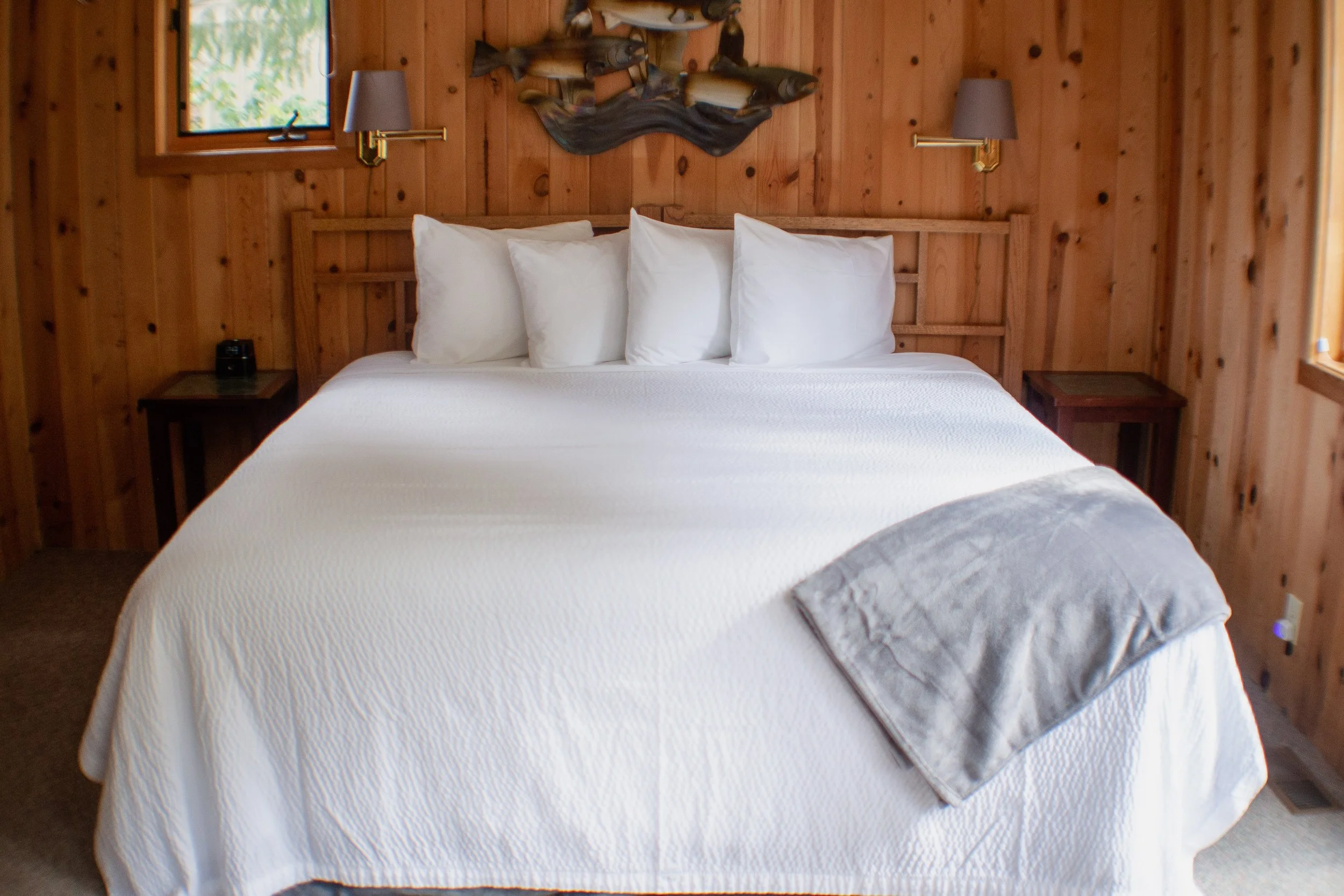 A bedroom with wooden walls and a large bed with white bedding and four white pillows. There are two small nightstands with lamps, a wall decoration above the bed, and two windows letting in natural light.