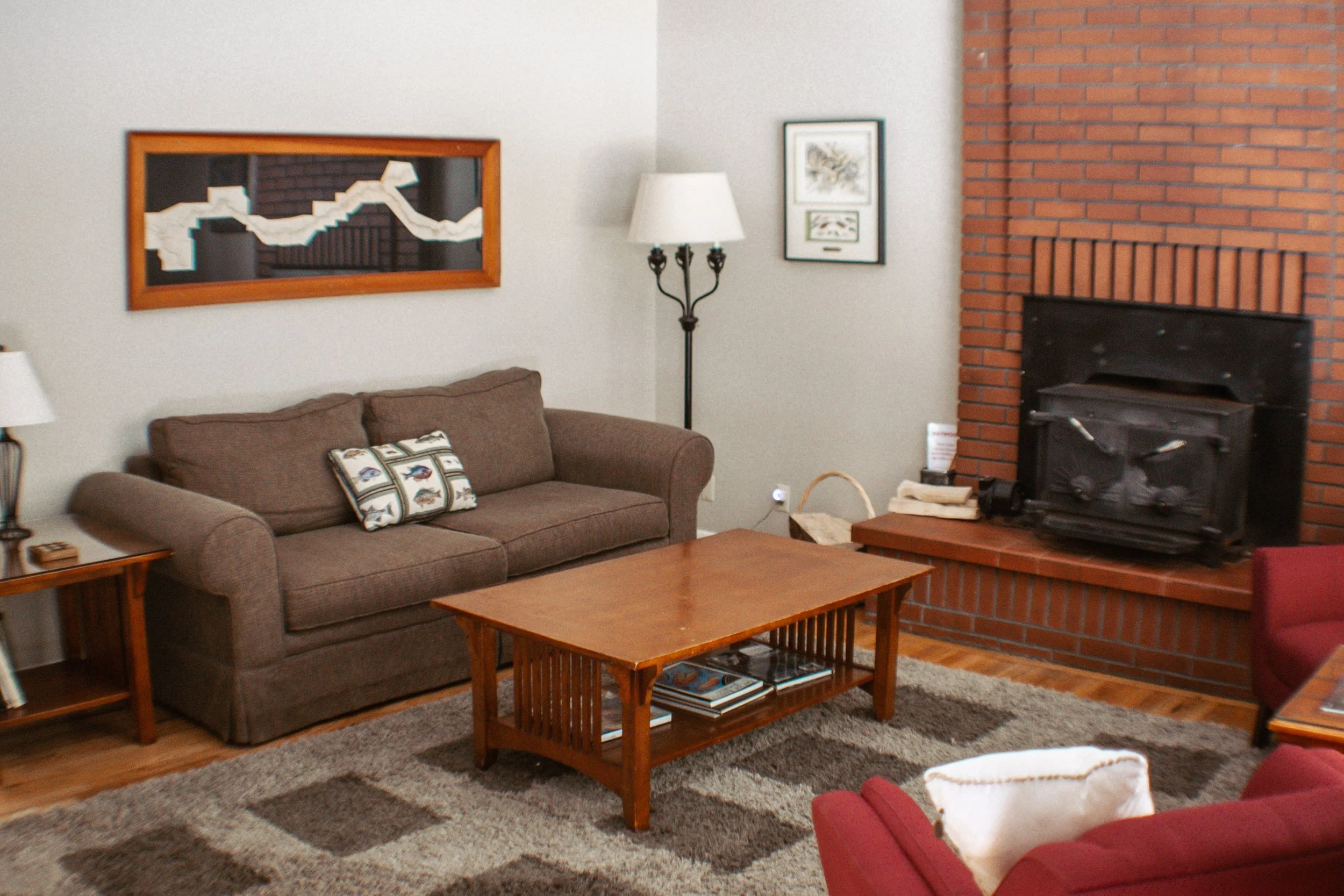 Living room with a brown sofa, a wooden coffee table, a red armchair, a brick fireplace, a floor lamp, framed pictures on the wall, and a patterned rug.