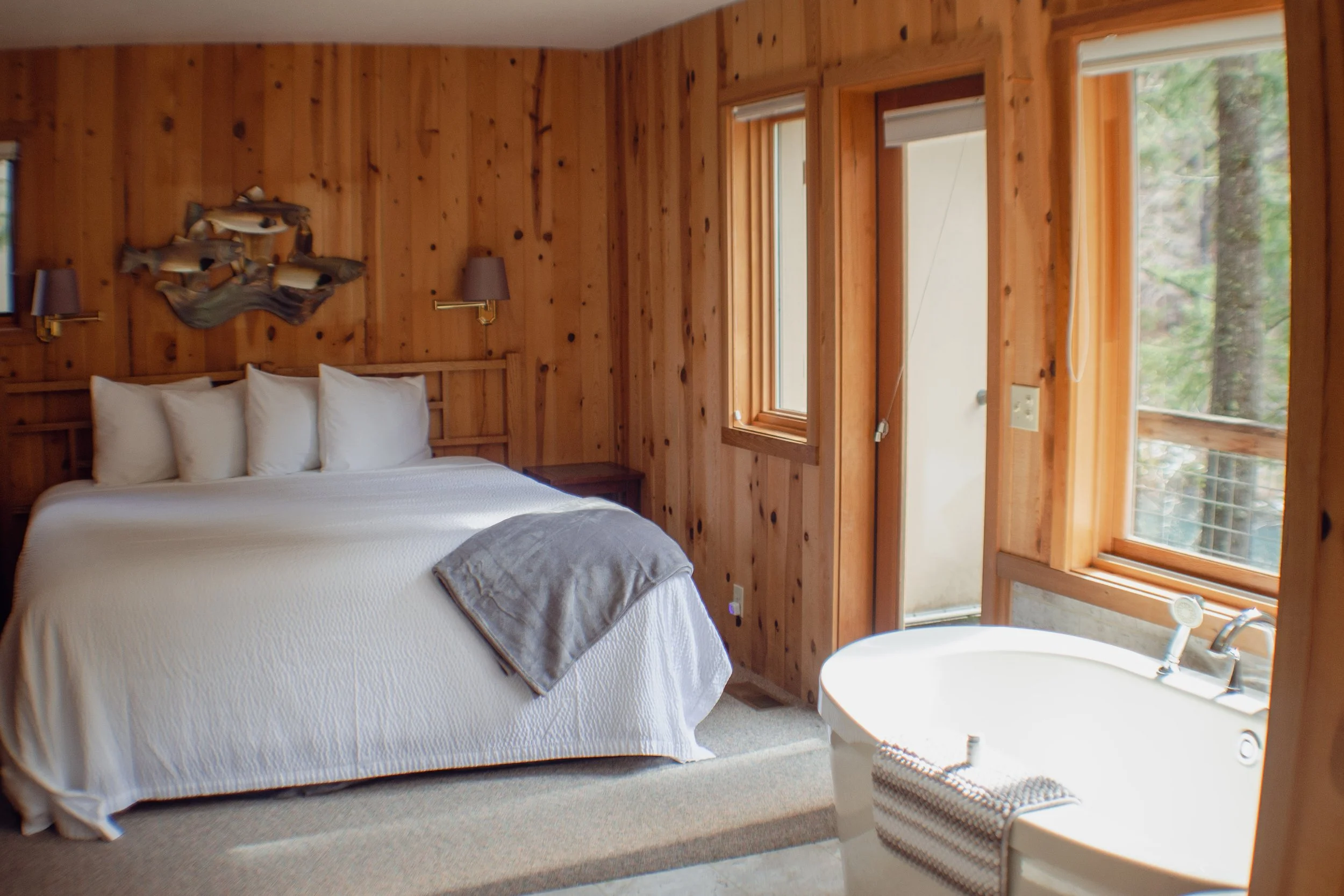 A cozy bedroom with wooden walls and a large bed with white bedding and multiple pillows, a blanket draped over the foot, and a bathtub near the window with a striped towel.