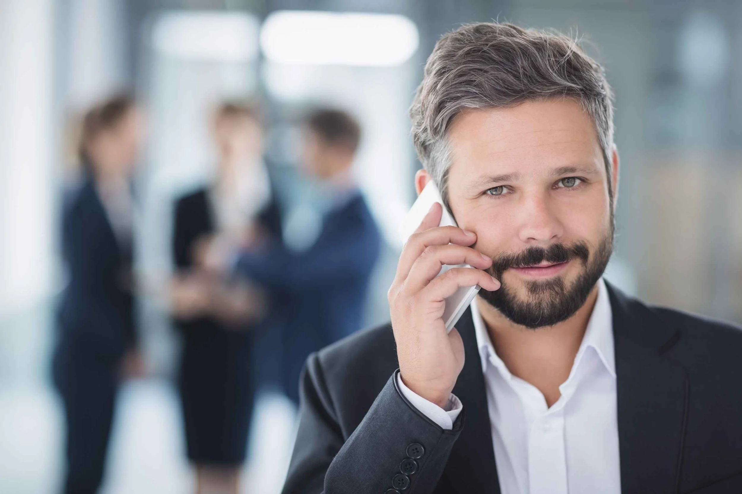 Businessman in a suit talking on a mobile phone, with three colleagues blurred in the background in a modern office setting.