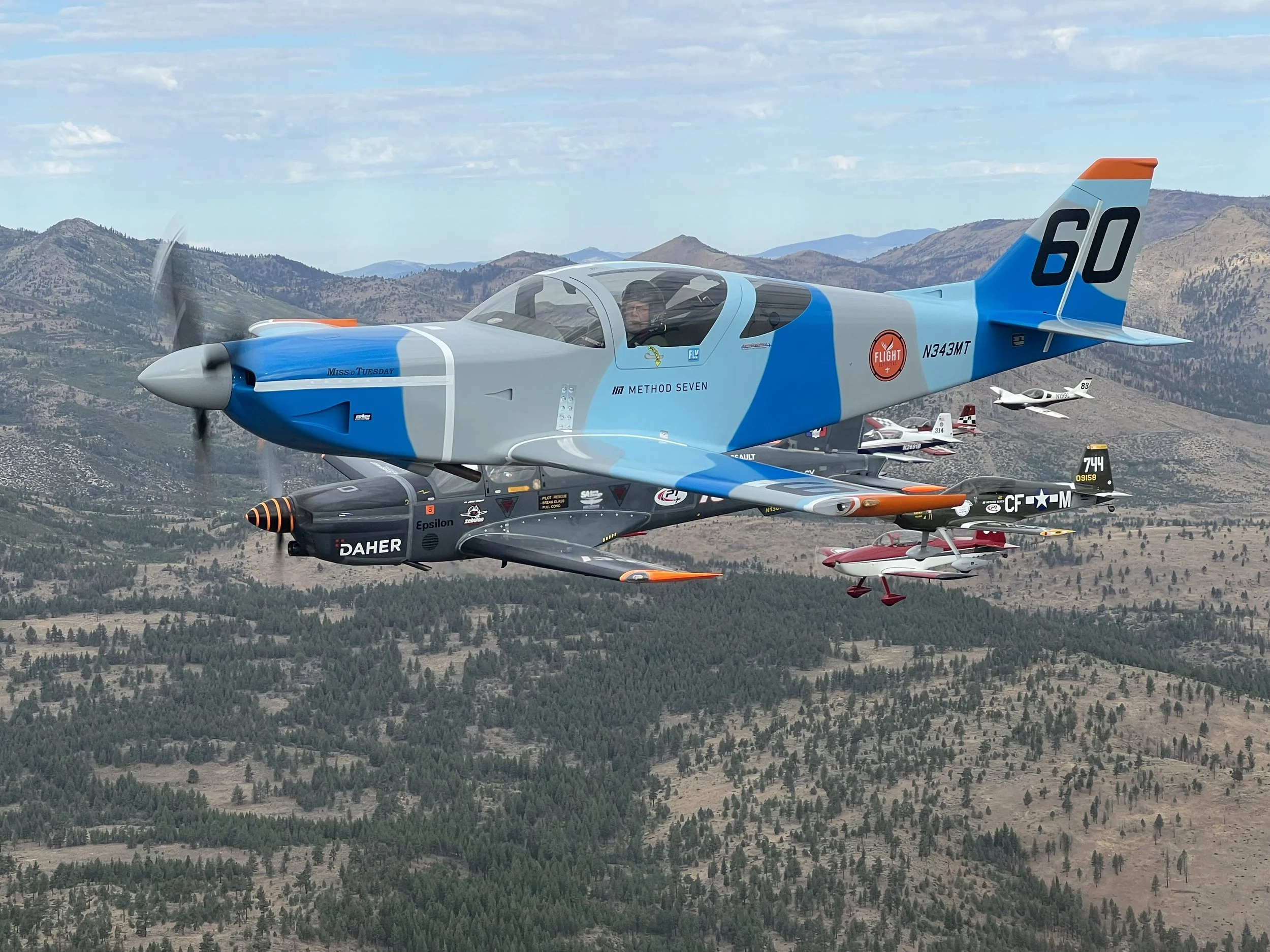 A formation of small aircraft flying over a mountainous landscape, led by a larger blue and gray jet with the number 60 on the tail, contrasted against a partly cloudy sky.