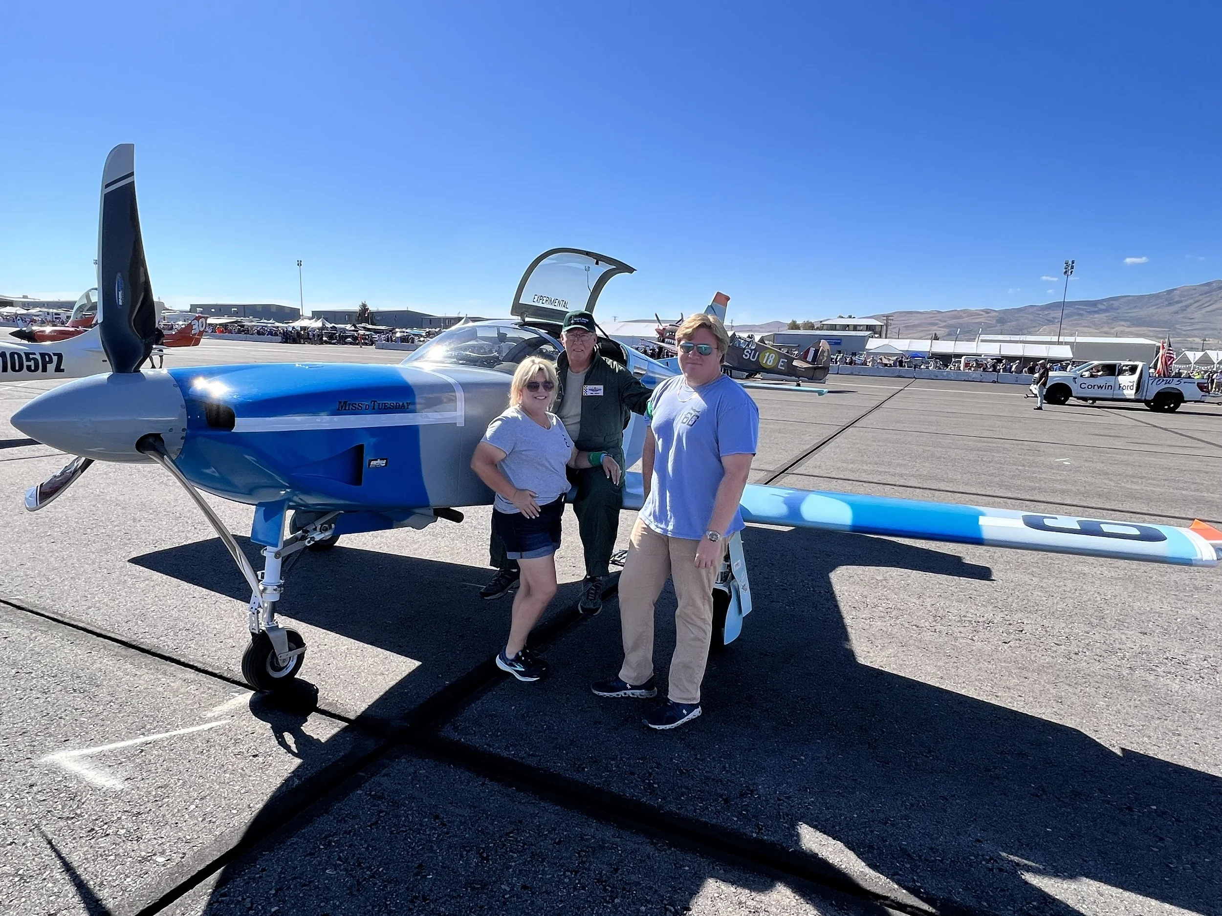 Three people standing next to a blue and white race plane on an airfield, with spectators and other planes in the background under a clear blue sky.