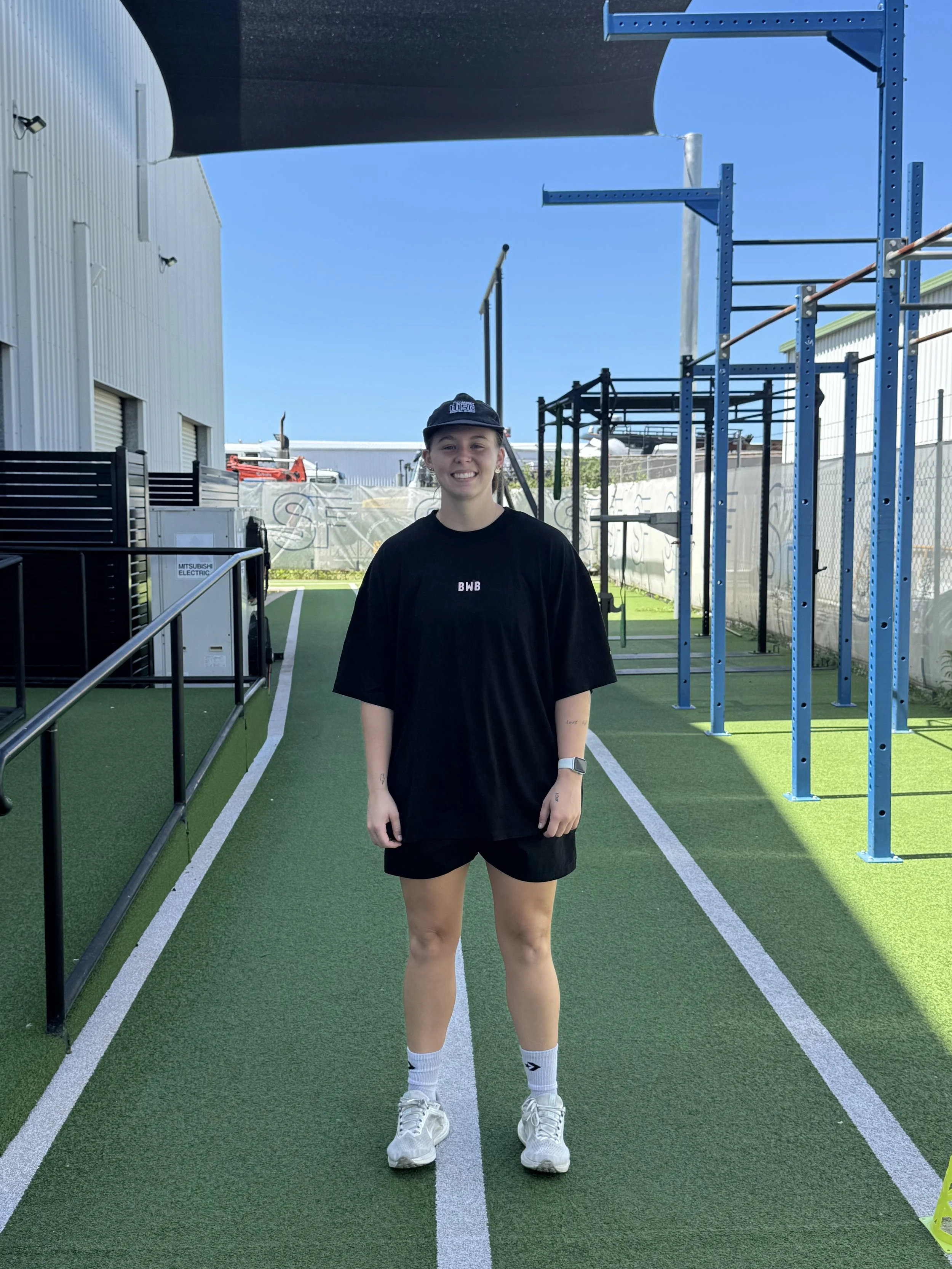 A young woman standing on an outdoor sports track, smiling, wearing a black t-shirt, black shorts, white sneakers, white socks, a black cap, and a smartwatch, with fitness equipment and a blue structure in the background.