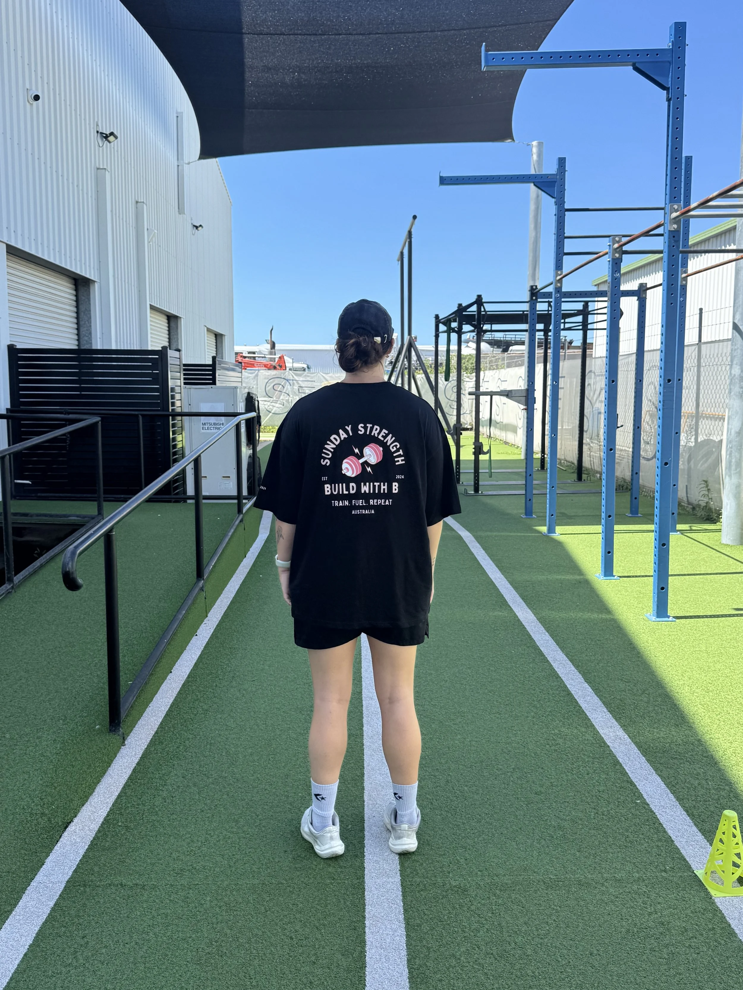 A person standing on a green track in an outdoor fitness area, facing away from the camera, wearing a black cap, black t-shirt with white and pink lettering, black shorts, white athletic shoes, and white socks, with blue equipment and a building in the background under a clear blue sky.