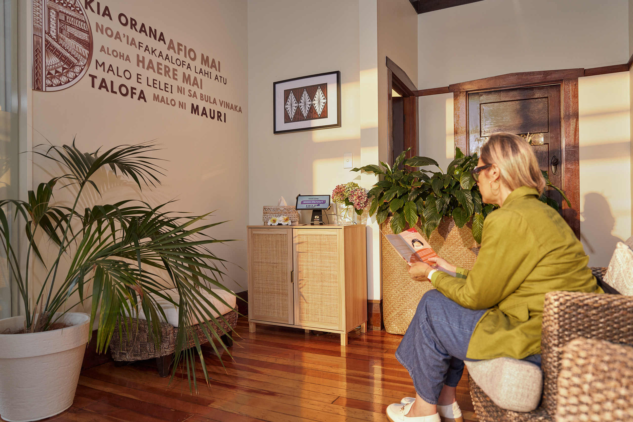 A woman with blonde hair wearing glasses, a green jacket, and a blue skirt is sitting on a chair reading a pamphlet in a waiting area decorated with plants and framed artwork. The wall has text in a foreign language and the floor is wooden.