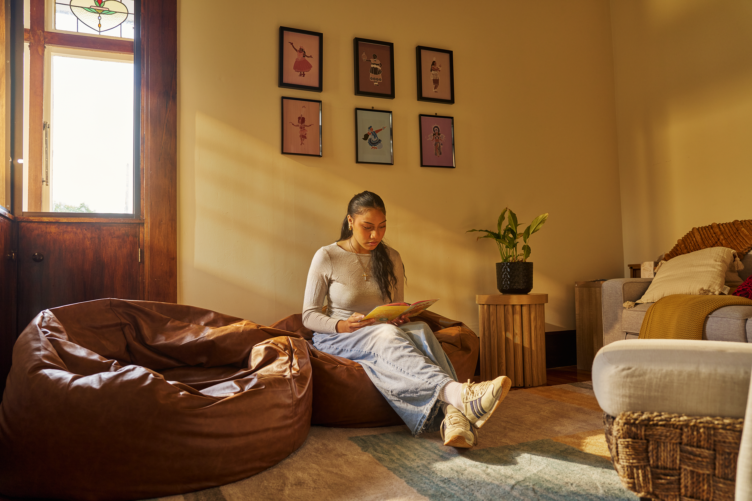 A young woman with long dark hair, dressed in a gray long sleeve shirt and light gray pants, sits on a large brown leather couch in a cozy living room, reading a magazine. The room has a warm yellow wall decorated with six framed pictures of dancers in colorful costumes. There is a wooden side table with a potted plant next to her and a bed with a beige headboard and pillows in the background, with soft natural light coming through a window.