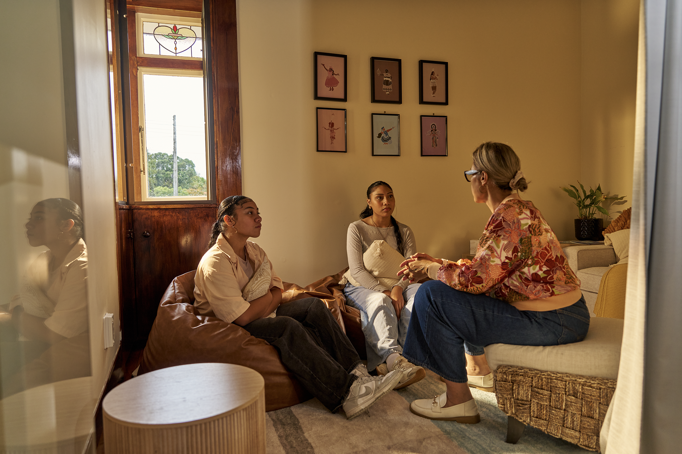 Four people, three women and one woman standing, are having a conversation in a cozy living room. The room has a window with wooden trim, framed art on the wall, a green plant, a lamp, and a patterned pillow on a sofa.