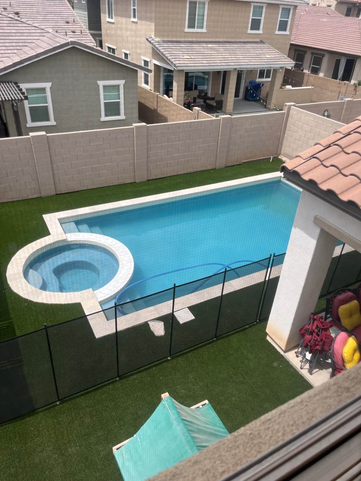 A backyard view with a swimming pool and hot tub surrounded by artificial grass, enclosed by a black safety fence, and adjacent to a house with outdoor seating visible on a covered patio.