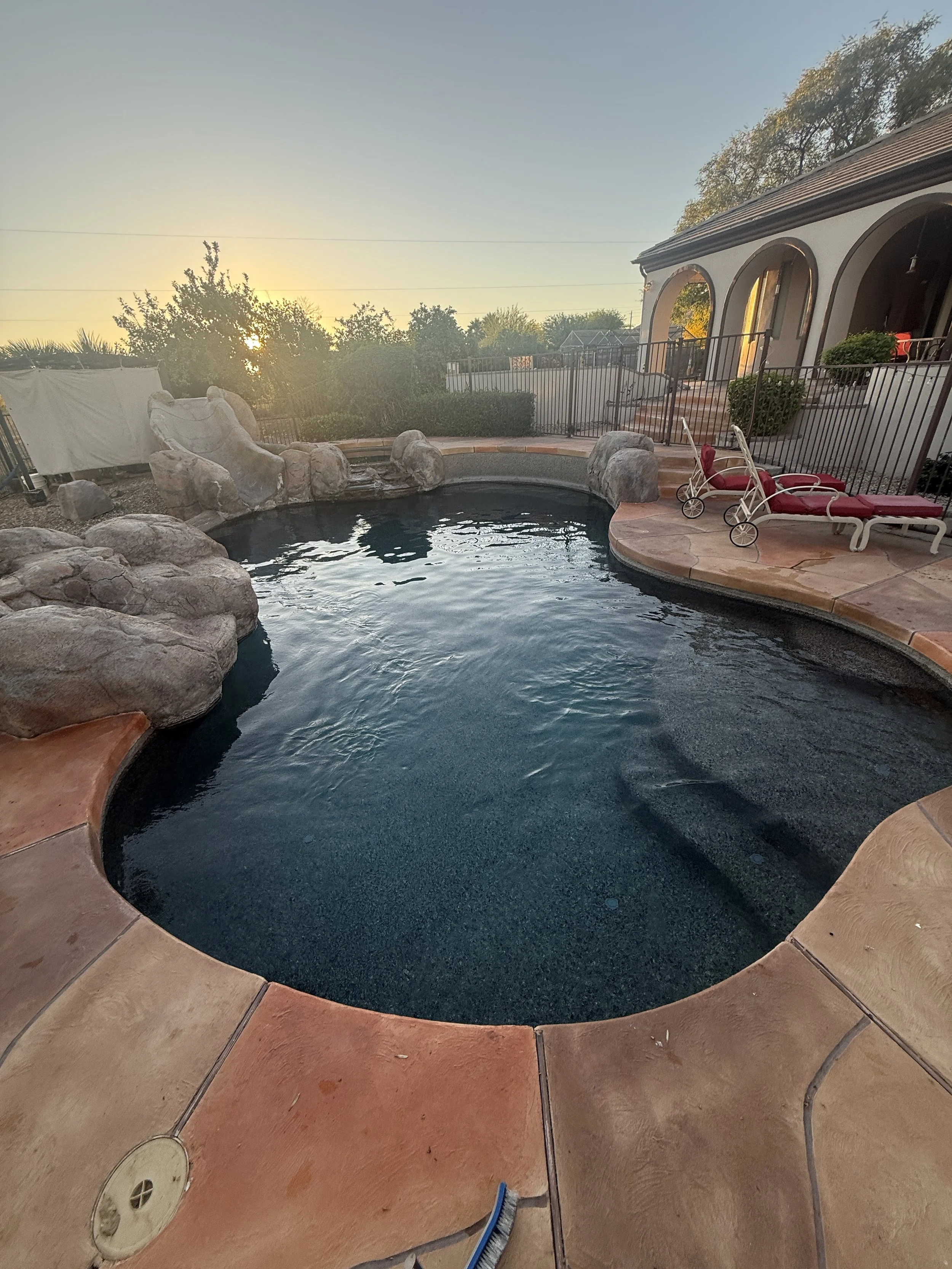 Private backyard with a small swimming pool, lounge chairs with red cushions, decorative rocks around the pool, green bushes, and a house with arched windows and a fence, during sunset.
