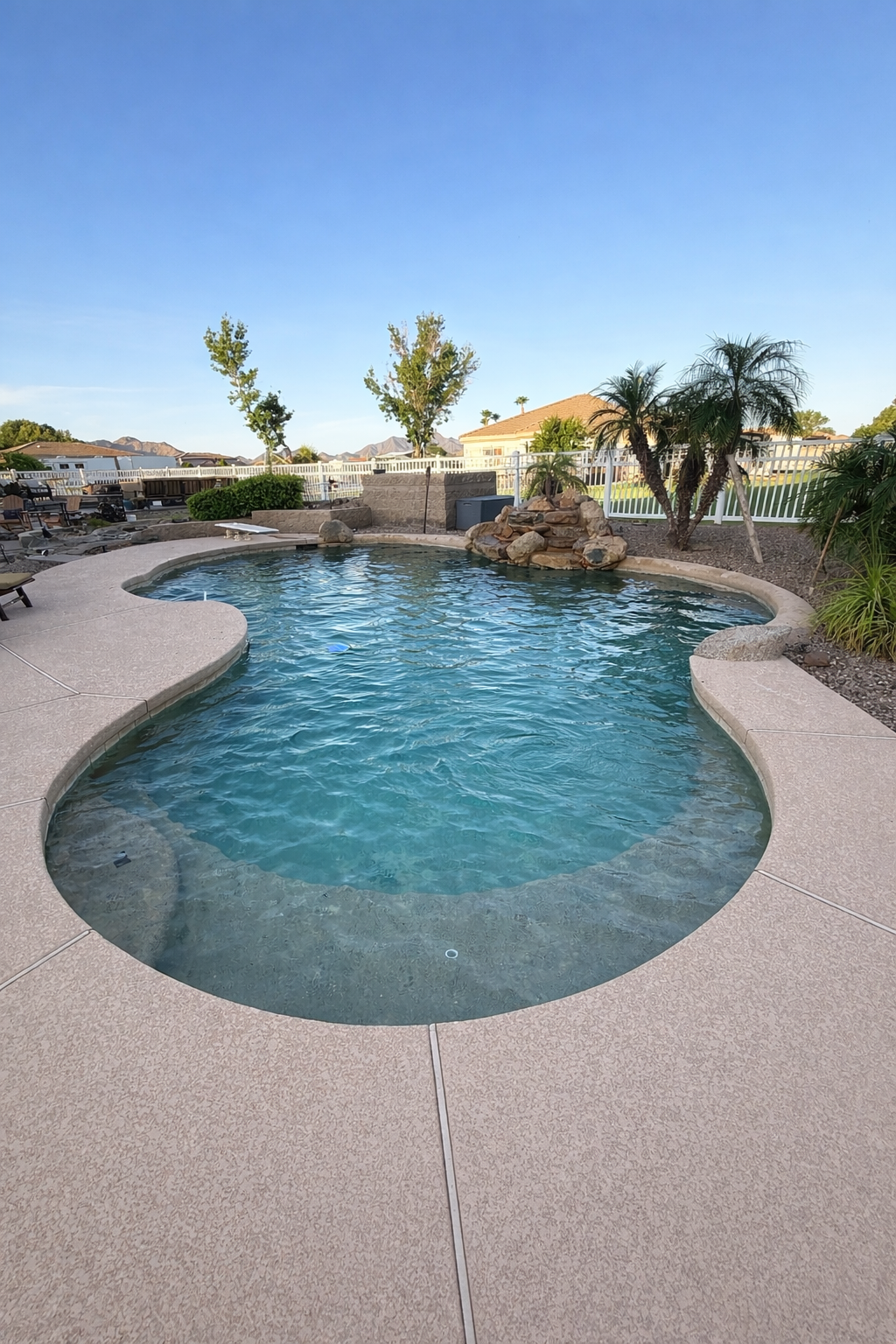 A backyard swimming pool with a rounded shape, surrounded by a textured pink concrete deck, with palm trees and other greenery in the background, under a clear blue sky.