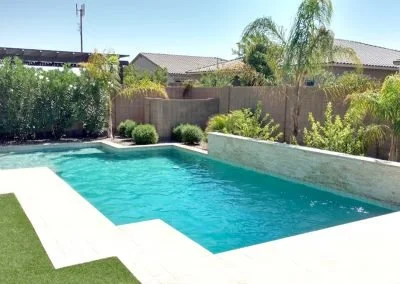 Backyard swimming pool with clear blue water, surrounded by a wooden fence and lush shrubbery, under a sunny sky.