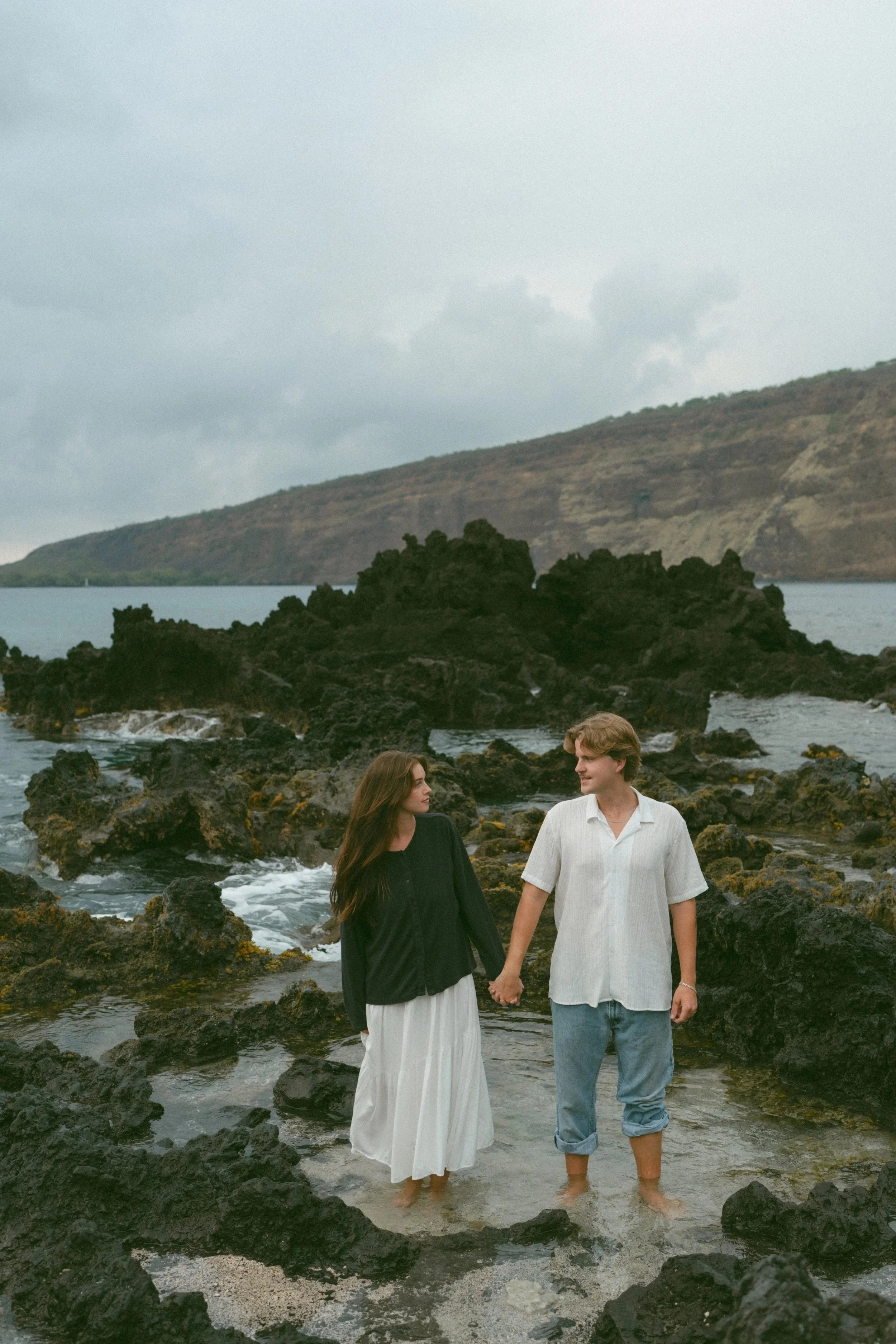 A young couple holding hands and walking on a rocky shoreline with water and hills in the background, overcast sky.