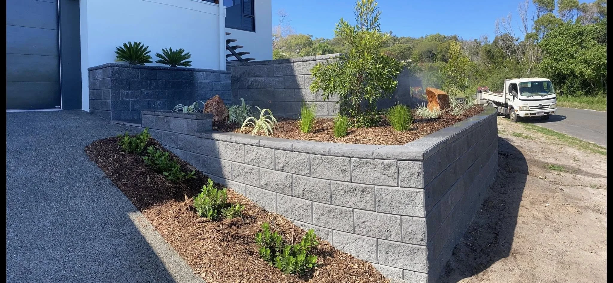 A newly built gray brick retaining wall surrounds a landscaped garden bed with plants and shrubs. A driveway is adjacent on one side, and a white truck is on the street in the background, with trees and a clear blue sky overhead.