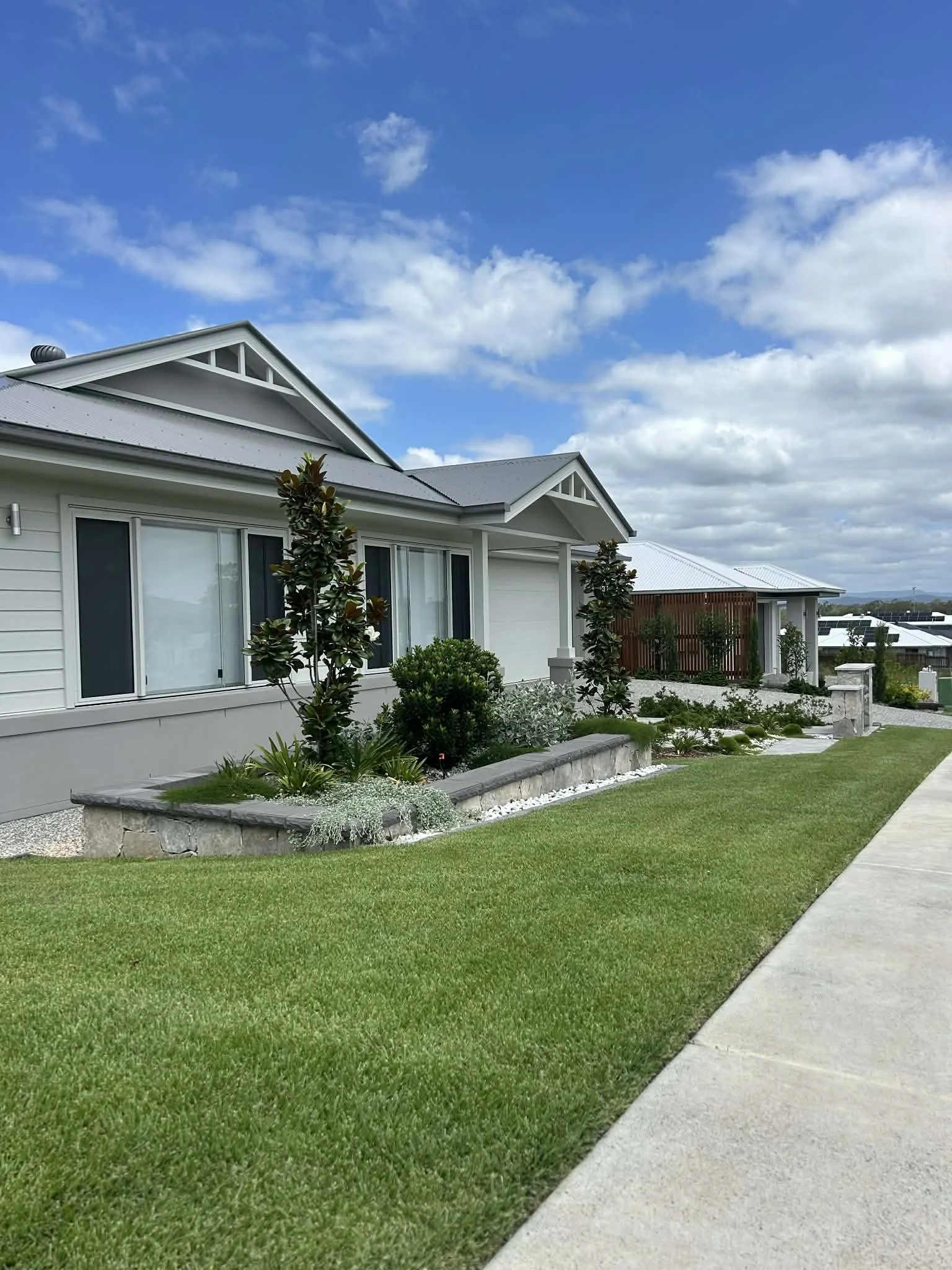 A modern house with white siding and a gray metal roof, surrounded by a neatly manicured lawn and landscaped garden beds with various shrubs and small trees, under a partly cloudy blue sky.