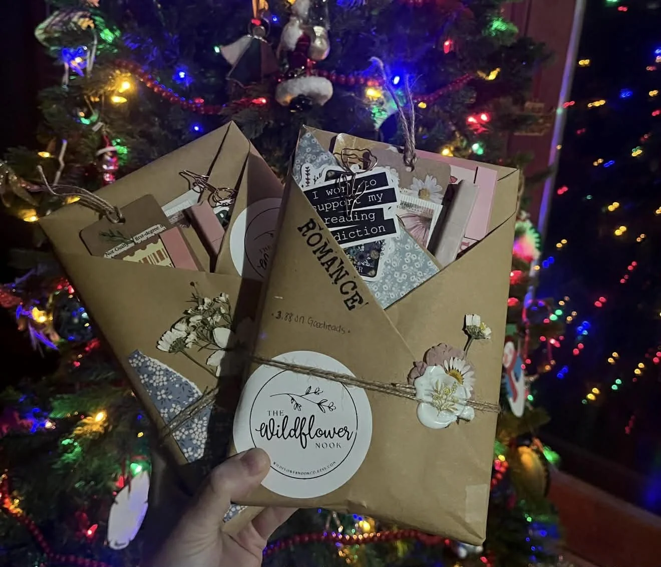 Hand holding a wrapped gift in front of a decorated Christmas tree with colorful lights and ornaments.