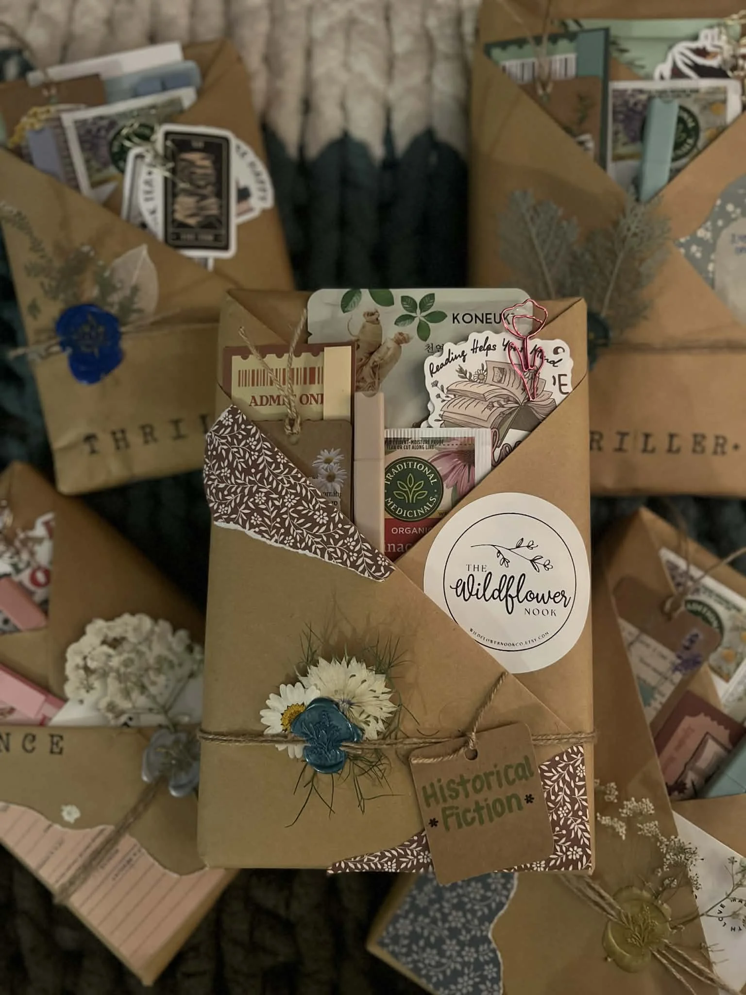 A gift box wrapped in brown paper with floral decorations, containing various books and booklets, labeled 'The Wildflower Book,' with a tag marked 'Historical Fiction,' and adorned with a flower and wax seal. Multiple similar wrapped gift boxes are visible in the background.
