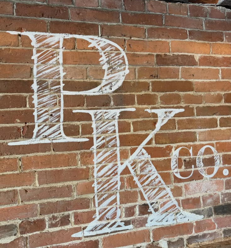White hand-drawn logo or sign on a brick wall with large stylized letters 'PK' and smaller text '.co'