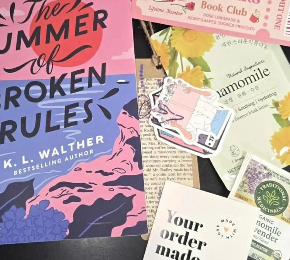Collection of various postcards and booklets, including a pink book titled 'The Summer of Broken Rules', a pink Book Club card, a herbal tea packet labeled 'Chamomile', and other promotional cards, set against a dark background.